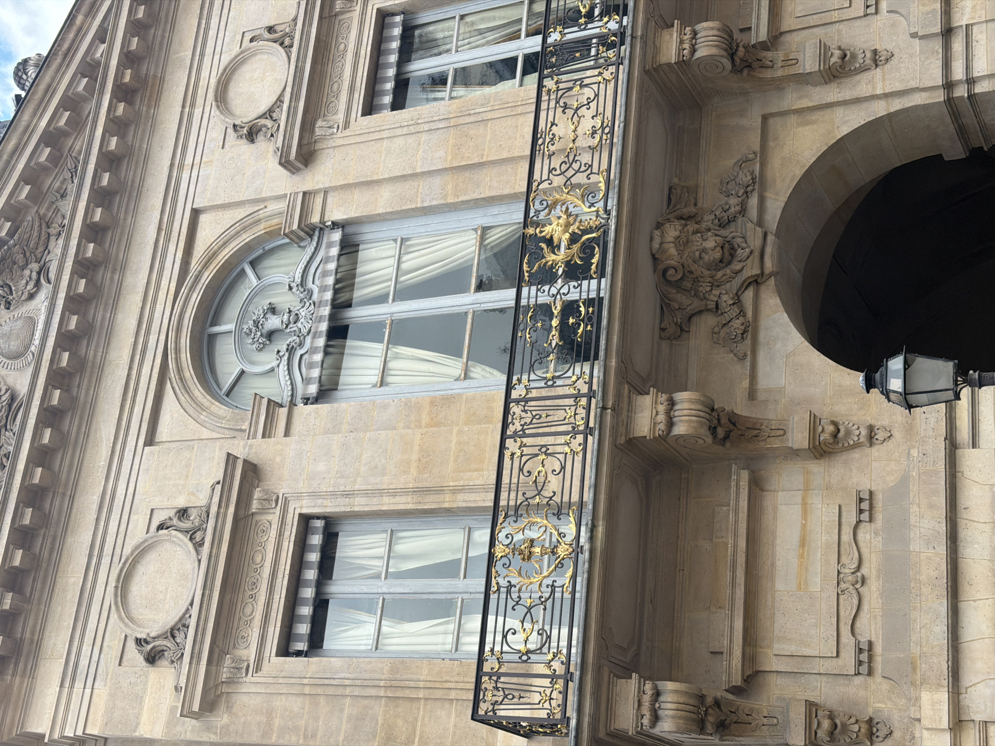A close-up, low-angle view captures a segment of an ornate building facade in Paris, France, exhibiting classical architectural grandeur. The structure is crafted from light-colored stone, adorned with intricate carvings and decorative mouldings across its multiple levels.
Several windows are visible: a prominent round window features a detailed central carving resembling an urn or vase, with spokes radiating outwards. Below and above it are rectangular windows, some showcasing white interior curtains, while others are partially covered by grey and white striped awnings.
A striking, narrow black wrought iron balcony or decorative grille extends vertically down the facade. It is exquisitely embellished with elaborate gold-painted scrollwork and flourishes, forming a central decorative element.
The stone facade is rich in intricate details, including carved oval plaques framed by decorative elements, classical corbels, and detailed mouldings. On the right side, a substantial carving of a lion's head with a flowing mane is visible, positioned near the opening of a dark stone archway, possibly an entrance or passage. A multi-faceted street lantern with a dark frame hangs above this archway.
A small patch of overcast or cloudy sky is visible at the top left, indicating daytime conditions. There are no people or visible text in the image, which focuses solely on the architectural craftsmanship and decorative artistry of the historic Parisian building.