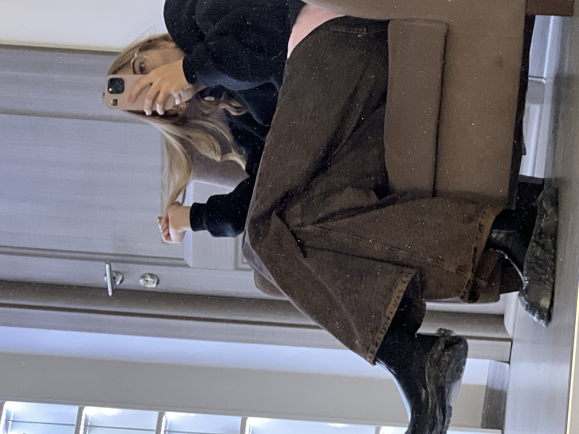The image displays a mirror reflection of a female subject seated in a brown upholstered armchair indoors. She is captured from the waist down, with her upper torso angled slightly right. The subject holds a light pink smartphone in her right hand, obstructing the majority of her face, and simultaneously lifts a section of her long blonde hair with her left hand. She wears clear-rimmed glasses, a dark long-sleeved top over a light pink garment visible at the waist, dark brown wide-leg trousers, and black chunky-soled boots. In the background reflection, a light-colored interior door with a silver handle and lock is visible to the right, and a multi-paneled structure, possibly a window or decorative glass block wall, is present to the left. The floor appears light-colored. The environment suggests an indoor setting, possibly a residence or private lounge. Numerous small, bright specks, consistent with dust particles on the reflective surface or camera sensor artifacts, are visible across the image. The precise location within Krasnoyarsk, Russia, cannot be determined from the visual information provided.