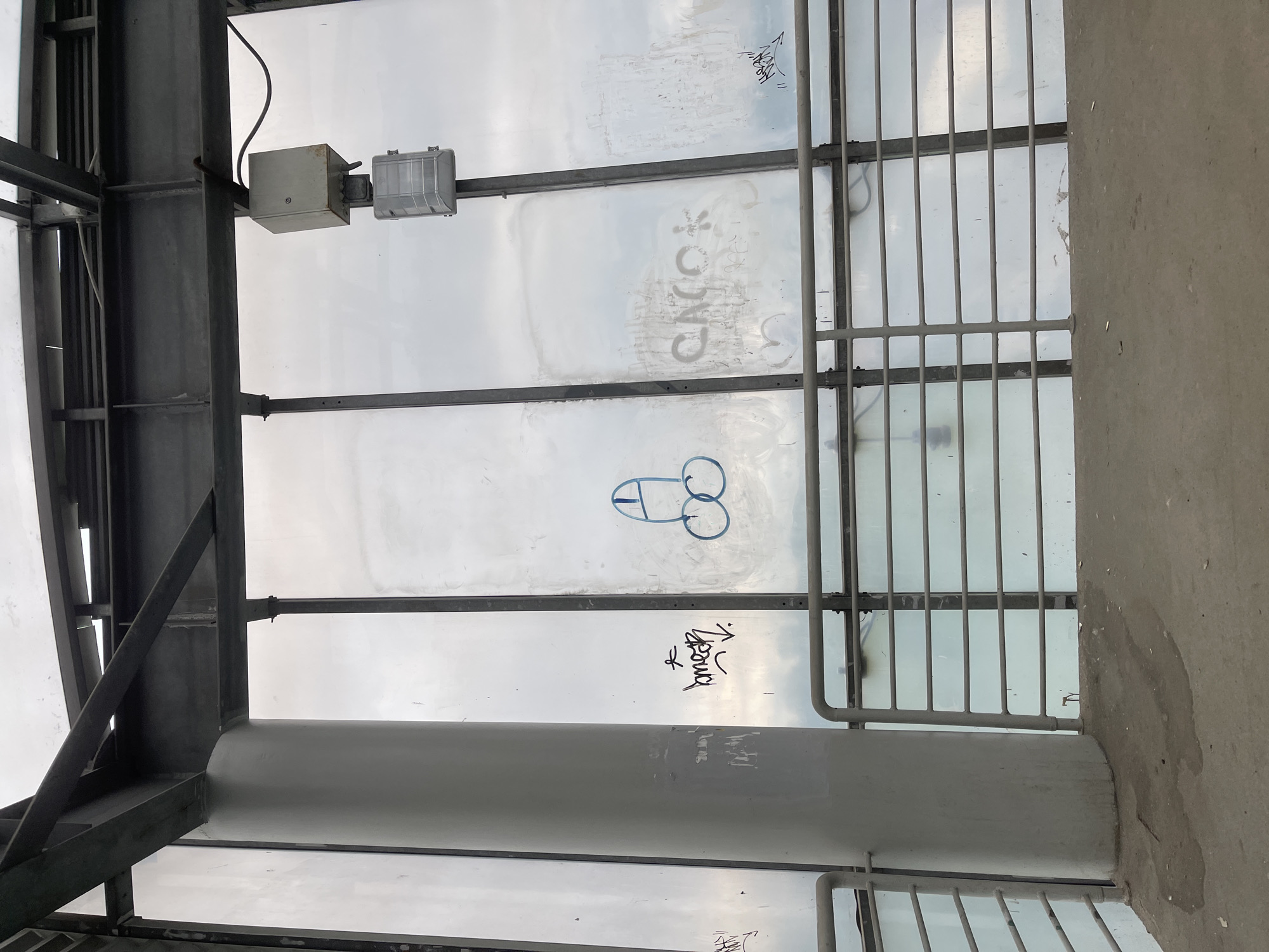 The image shows an interior structure with a view of the sky, possibly a pedestrian overpass or bus shelter. Graffiti is visible on the transparent panel, including a drawing of male genitalia and illegible text. The structure is made of metal and concrete. Location specifics for Razvilka, Russia, cannot be determined from the image. No people are visible.