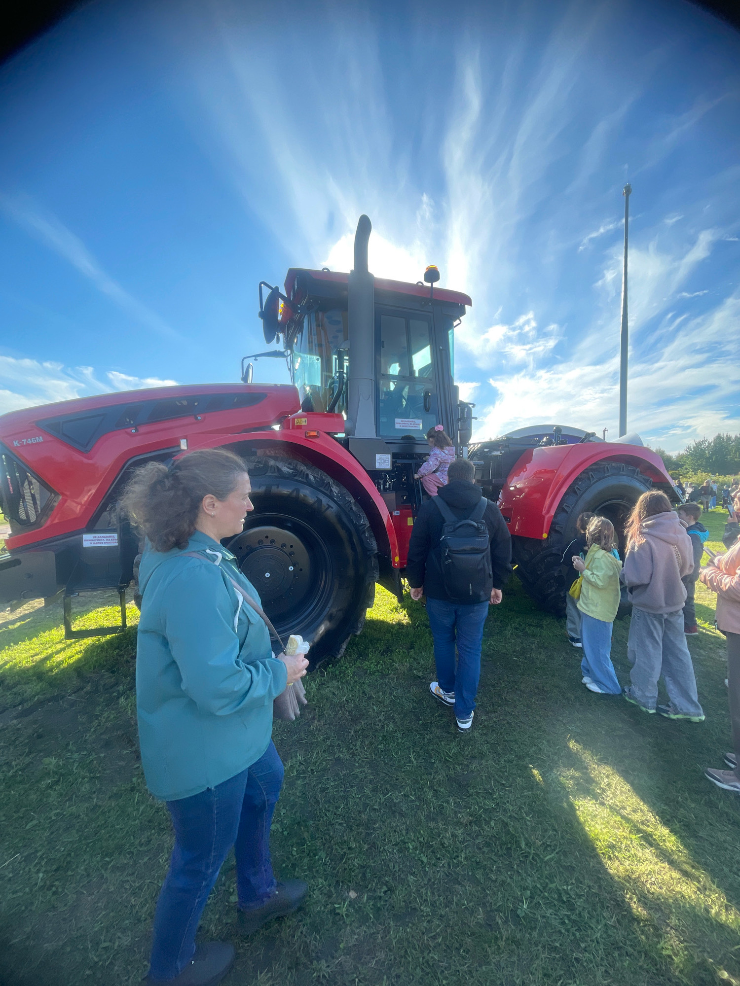 The image depicts a bright, sunny day with a clear blue sky filled with wispy white clouds. A large, red tractor is the central focus, with its front and side visible. Several people are gathered around the tractor, some appearing to be examining it, while others are walking by or standing in small groups. A woman in a turquoise jacket is in the foreground, facing away from the camera and towards the tractor. A young child is climbing into the tractor's cab. The scene appears to be outdoors on a grassy area, possibly at an agricultural exhibition or fair. The overall atmosphere is casual and family-friendly. There is no discernible information in the image to suggest a specific location in Saint Petersburg, Russia.