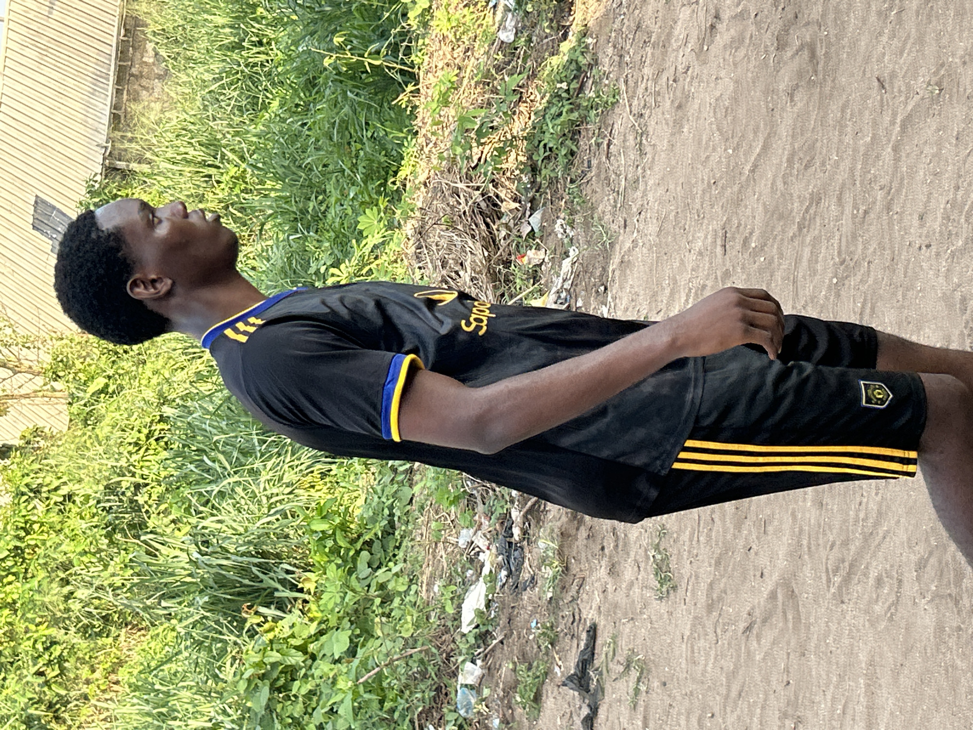 A young Black man is standing on a dirt path in Ihiagwa, Nigeria. He is wearing a black soccer jersey with yellow accents on the collar and sleeves. The jersey has a sponsor logo on the front. He is also wearing black soccer shorts with yellow stripes and a team logo on the right thigh. The path is surrounded by vegetation. There is a corrugated metal building visible in the background.