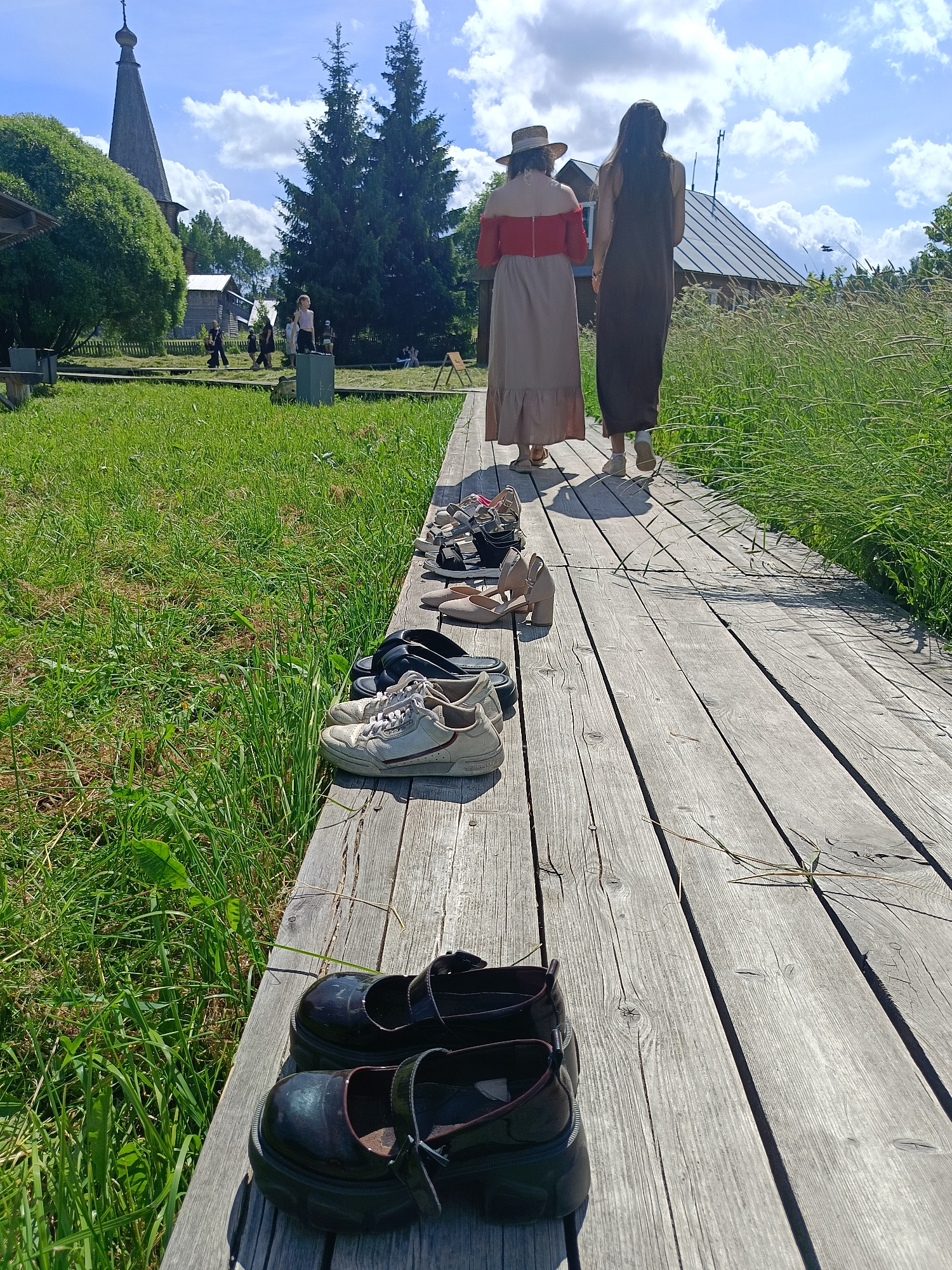 Two women, viewed from behind, walk along a wooden boardwalk in a rural, outdoor setting under a partly cloudy sky. The woman in the foreground wears a wide-brimmed hat and a light-colored, floor-length dress with an off-the-shoulder red top. The woman behind her wears a dark, floor-length dress. A variety of shoes are lined up on the left side of the boardwalk, including black platform Mary Janes, white sneakers, black sandals, and beige heeled sandals. The boardwalk is flanked by a grassy area on the left and a field of tall grass on the right. In the background, several wooden buildings, including one with a tall, pointed spire, and some pine trees are visible. The overall impression is a casual, summer day in a village or museum setting, possibly in Popovka, Russia, given the architectural style of the buildings.