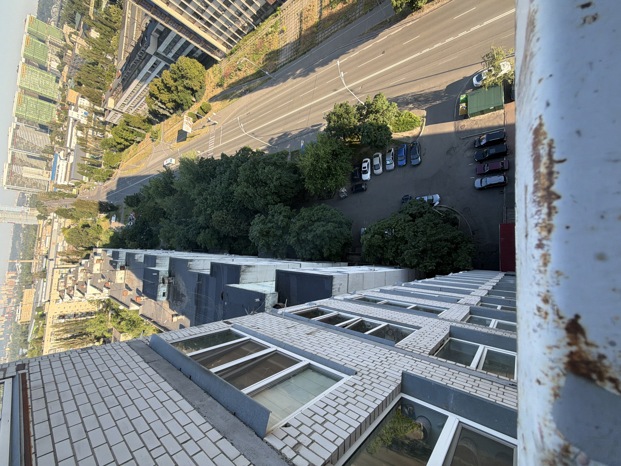 The aerial view shows a street in Kyiv, Ukraine, with a multi-story brick building in the foreground. The building has many windows and appears to be an apartment complex or office building. A road runs parallel to the building, with several cars parked along its side. Trees line the street, and in the distance, a cityscape with tall buildings is visible. The sky is clear and the sun is shining, casting shadows across the scene.