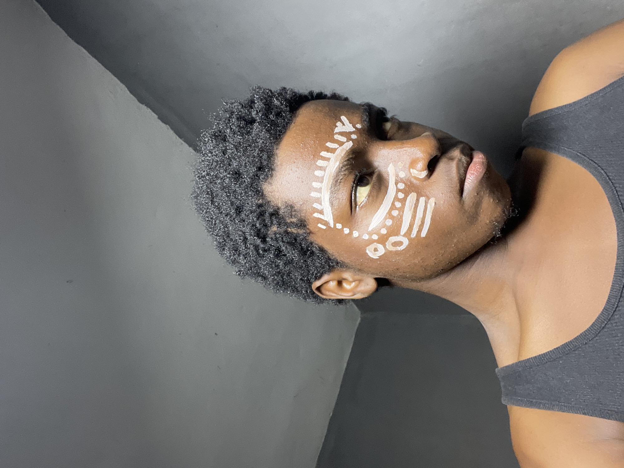 The image shows a close-up of a young person with dark skin and short, curly black hair. They are wearing a dark gray tank top. Their face is adorned with white face paint, featuring geometric patterns and dots around the eyes and on the cheeks. The person is looking to the right of the frame with their head slightly tilted. The background is a plain, dark gray wall, suggesting an indoor setting. There is no discernible indication of the location being in Nkpor, Nigeria; this is a generic indoor portrait.