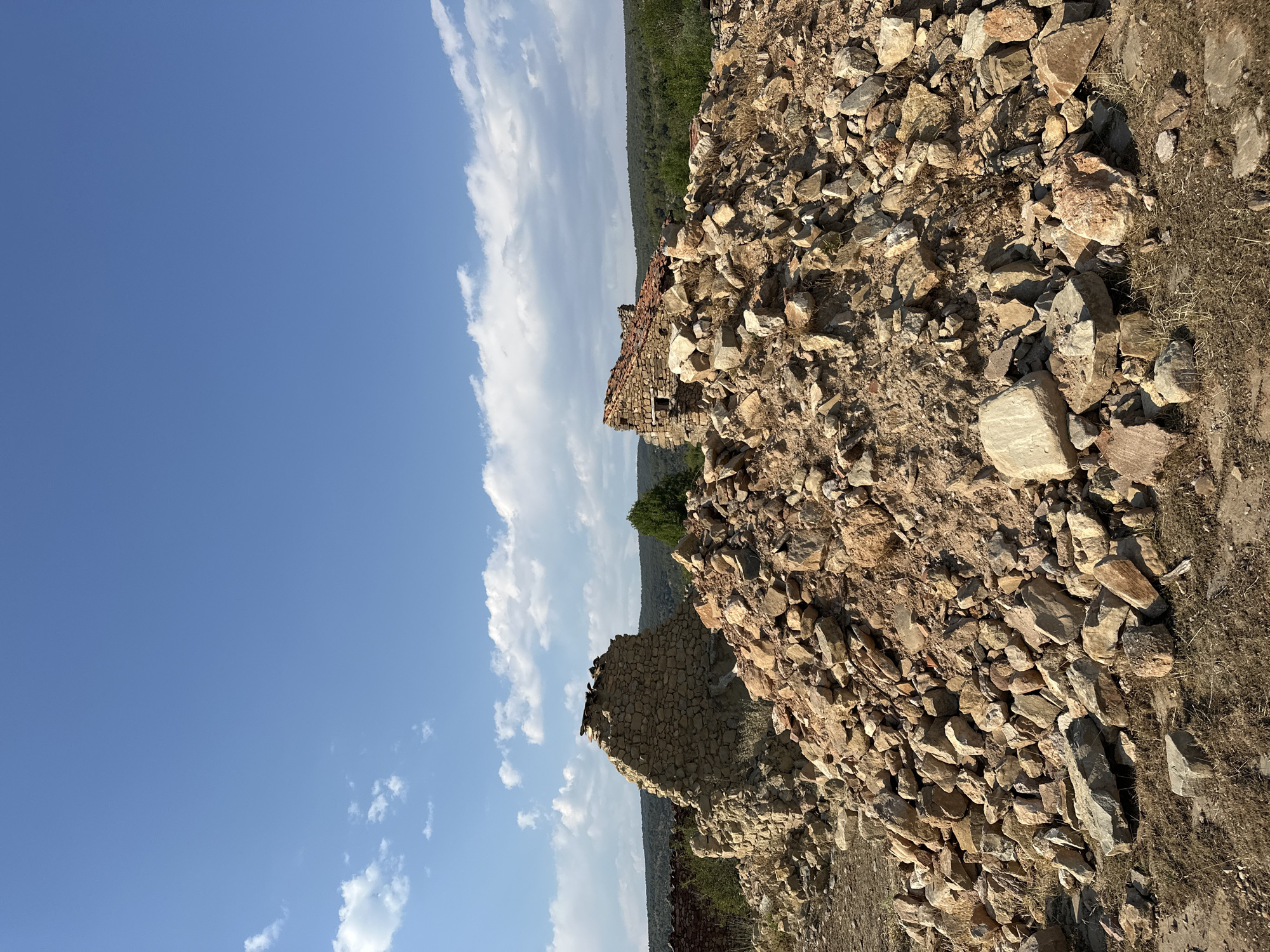 A low-angle, outdoor, full shot captures the ruins of two stone structures against a bright blue sky dotted with white clouds. In the foreground, a large, disordered pile of rocks, varying in size and color from light tan to reddish-brown, dominates the lower half of the frame. Some dry grass and small weeds are visible amongst the stones. Behind this rubble pile, the partially collapsed remains of two stone buildings are visible. The structure on the left is a tall, thick wall of rough-hewn stones, with a section of weathered red roof tiles precariously balanced on top. To the right, another building fragment shows a similar stone construction with a steeply angled, partly intact red tile roof. Small windows or openings are visible in this structure. In the middle ground, a dense line of green trees and vegetation stretches across the horizon, suggesting a wooded area or hillside. A single, larger green tree stands prominently behind the rightmost ruin. In the far background, a distant, undulating line of hills fades into the background. The overall impression is one of decay and abandonment in a rural, possibly Mediterranean, landscape. The scene is likely located in Alpuente, Spain, given the architectural style and the characteristic landscape.