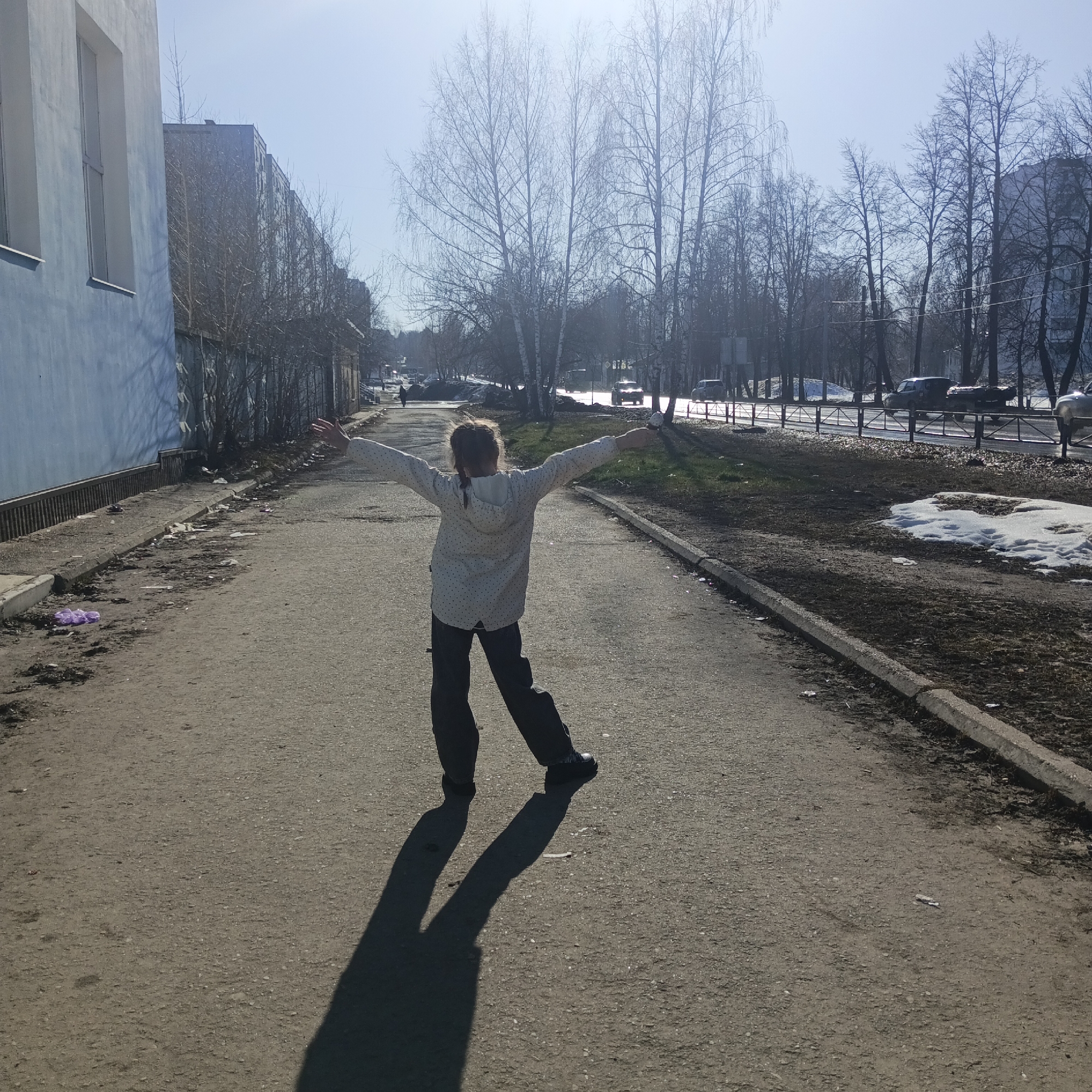 The image shows a girl standing on a paved road in the city of Chaikovsky, Russia. She has her arms outstretched, facing away from the camera. She wears a light-colored polka-dot jacket and dark pants. Her shadow stretches long on the road in front of her.
On the left side of the road, a light blue building with windows is visible. Litter is scattered along the edge of the road near the building. On the right side of the road, a strip of land is visible with patches of grass and melting snow. A metal barrier runs along the edge of the road, and cars are visible in the distance beyond it. Trees without leaves line the background. The sky is bright and clear.