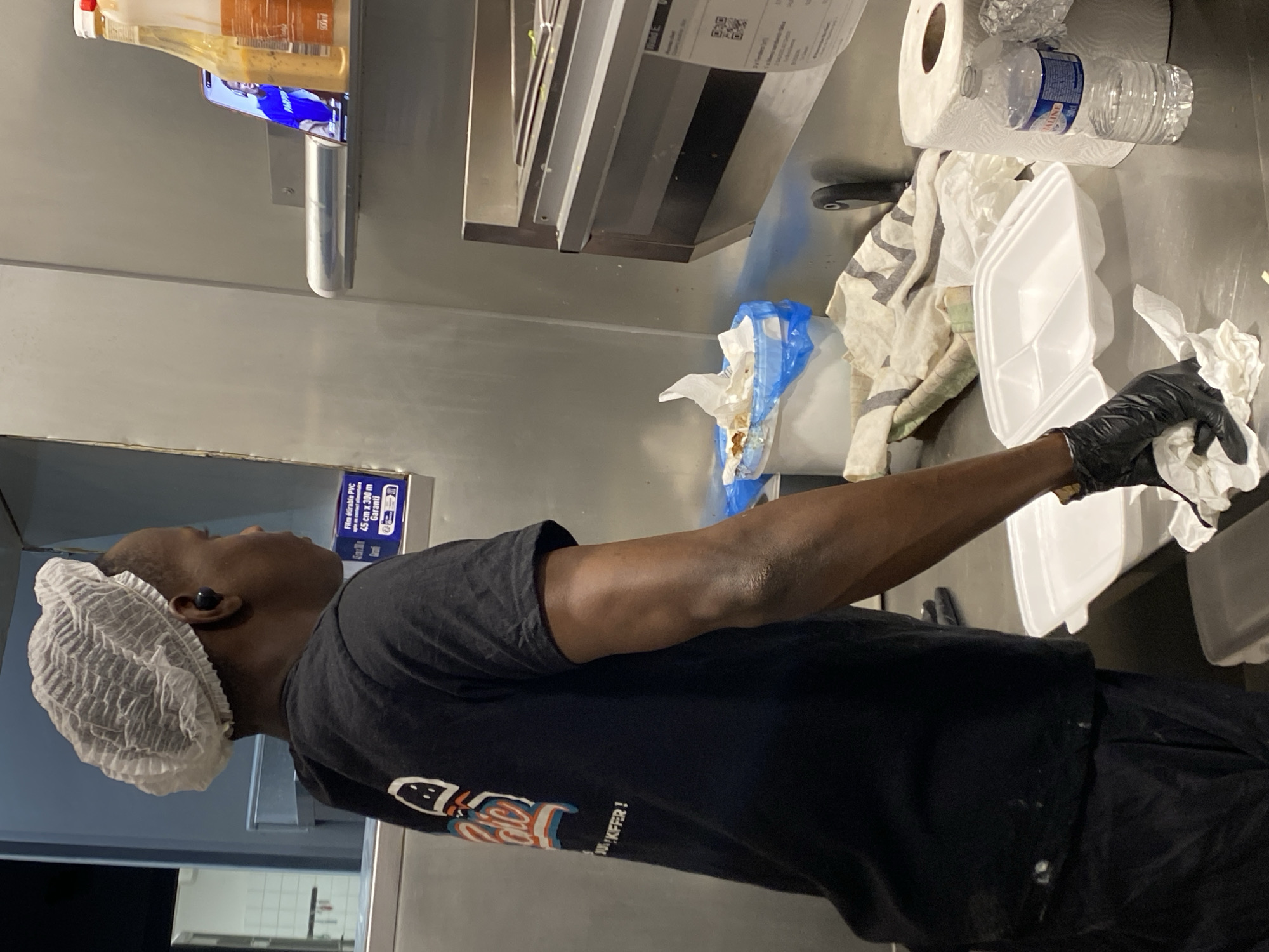 The image captures a person in a food preparation setting, likely a commercial kitchen, indicated by the stainless steel surfaces and professional equipment. The scene is illuminated by artificial light, suggesting it could be any time of day or night.
The primary subject is a person, likely male, with dark skin, visible from the chest up. They are wearing a white disposable hairnet and a black short-sleeved t-shirt with a graphic design; the text "KFFER!" is partially visible on the shirt. An earbud is placed in their right ear. The person's right arm is extended forward, and they are wearing a black glove on their hand, holding a crumpled white paper towel or tissue, seemingly wiping the inside of a white, multi-compartment disposable food container. Other black-gloved fingers are visible near the container.
On the metallic counter to the right, there's a jumble of items: several crumpled paper towels, a roll of paper towels, an empty plastic water bottle with "VALTALINE" clearly printed, a striped cloth (beige, white, grey), and more white disposable food containers. A blue plastic bag containing what appears to be food scraps or refuse is also present.
Above the counter, a shelf holds two bottles of condiments or sauces, one with an orange hue and a red cap. Beneath these bottles, a mobile phone is placed horizontally, displaying a screen with blue elements. Further to the left, on a shelf, there's a box of "Film étirable PVC 45 cm x 300 m". A receipt or order slip with a QR code is visible hanging near the top. The overall impression is one of an active, functional, and somewhat cluttered workspace.
