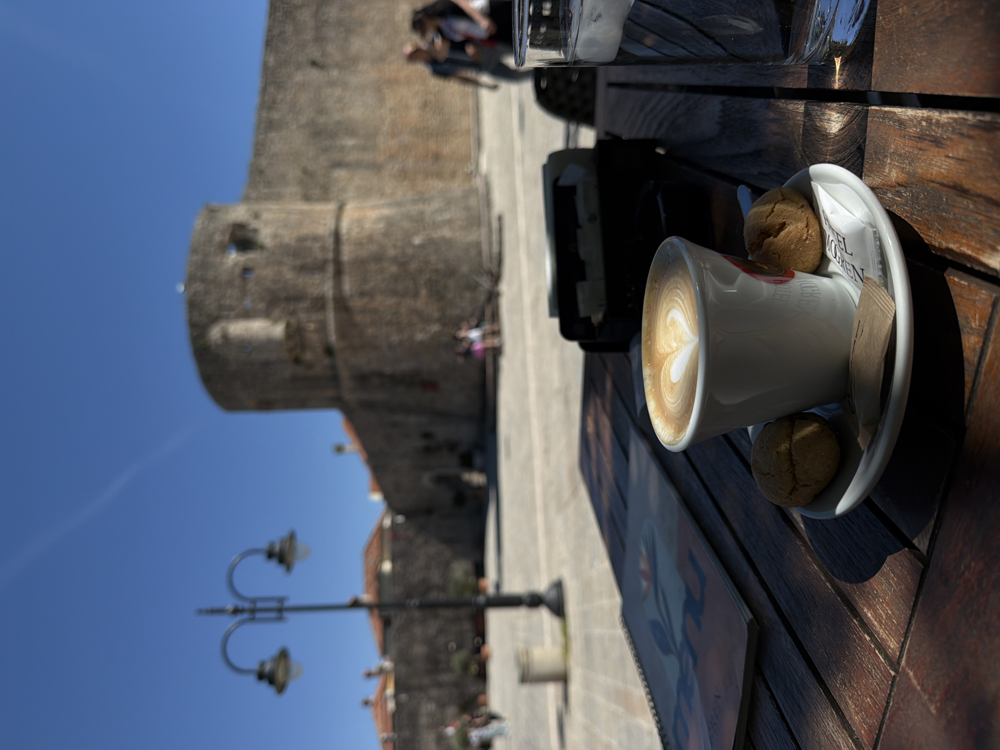The image depicts an outdoor cafe setting with a historical backdrop, likely in Budva, Montenegro, as suggested by the visible "HOTEL MOGREN" text on a sugar packet. The scene is brightly lit by the sun, indicating midday or early afternoon under a clear blue sky.
In the foreground, a white cup of coffee, possibly a latte or cappuccino, sits on a matching white saucer on a dark, slatted wooden table. The coffee features delicate heart-shaped latte art. Two small, golden-brown cookies are placed on the saucer, one prominent and another peeking from underneath the cup. Accompanying the coffee are a sugar packet, clearly displaying the text "HOTEL MOGREN", and a brown stirrer. Partially visible on the table are a clear glass, possibly containing water, a dark rectangular object (perhaps a phone or camera), and a menu or brochure with an illustrated design. Strong shadows are cast across the wooden table, emphasizing the sunny conditions.
The background showcases the impressive architecture of an old European town. A massive, light-colored stone wall, resembling an ancient fortress or city wall, dominates the left side of the background. Further back, traditional street lamps with ornate designs and old buildings with terracotta roofs can be seen. Several blurred figures are scattered in the mid-ground, suggesting people walking or enjoying the bustling atmosphere of the historic location. The overall impression is one of leisure and enjoyment in a picturesque historical setting.