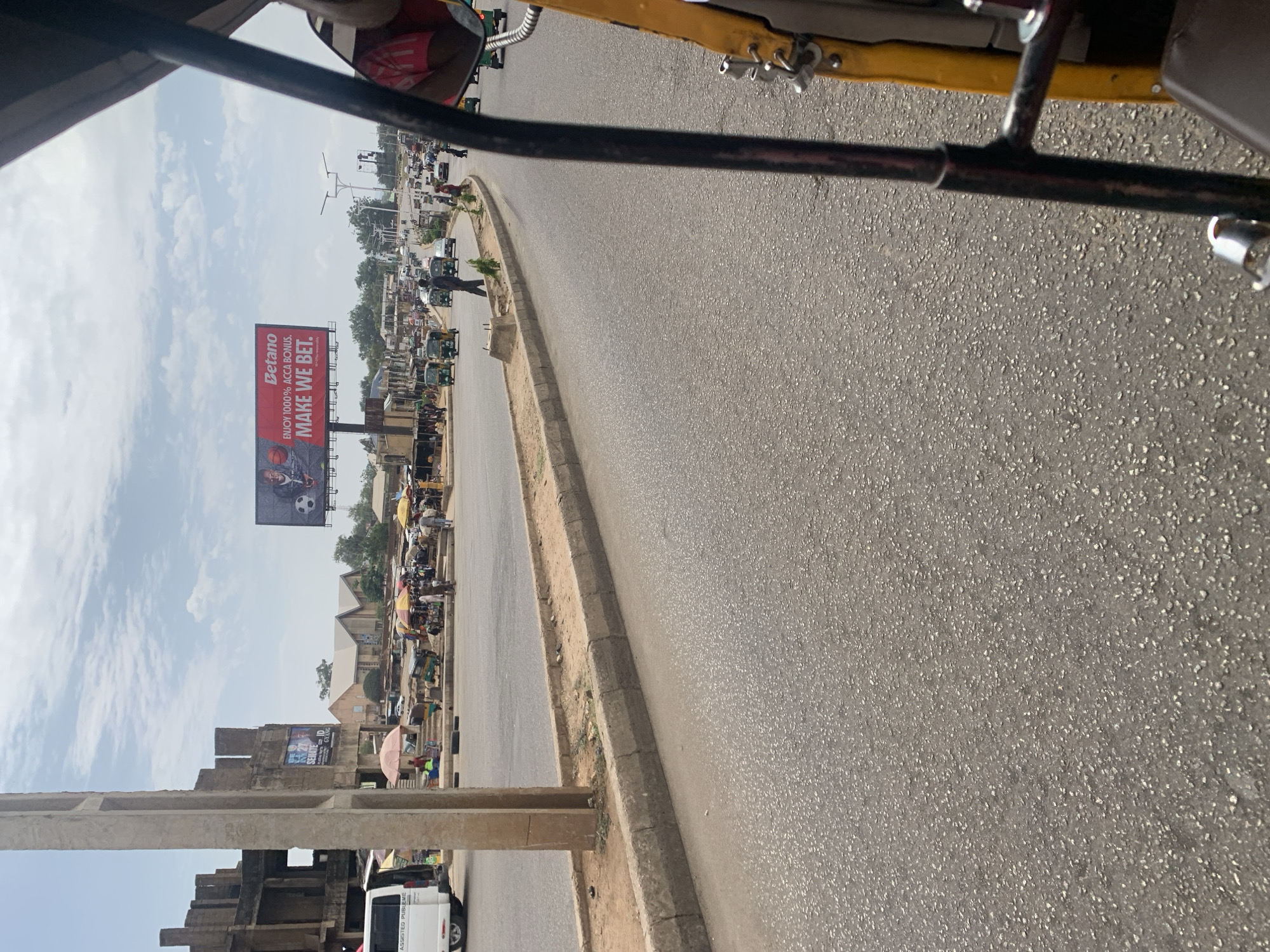 The image shows a street scene in Kabong, Nigeria, captured from a moving vehicle, likely an auto-rickshaw, as part of its frame is visible. The road is asphalt and stretches into the distance, featuring a concrete divider on the left side. A Betano billboard is visible on the left, featuring a soccer player and the slogan "ENJOY 100% ACCA BONUS. MAKE WE BET.". The skyline includes buildings and trees, and numerous auto-rickshaws and people are present along the road. There is a construction site with scaffolding and a white van parked nearby. Street vendors are visible under colorful umbrellas.