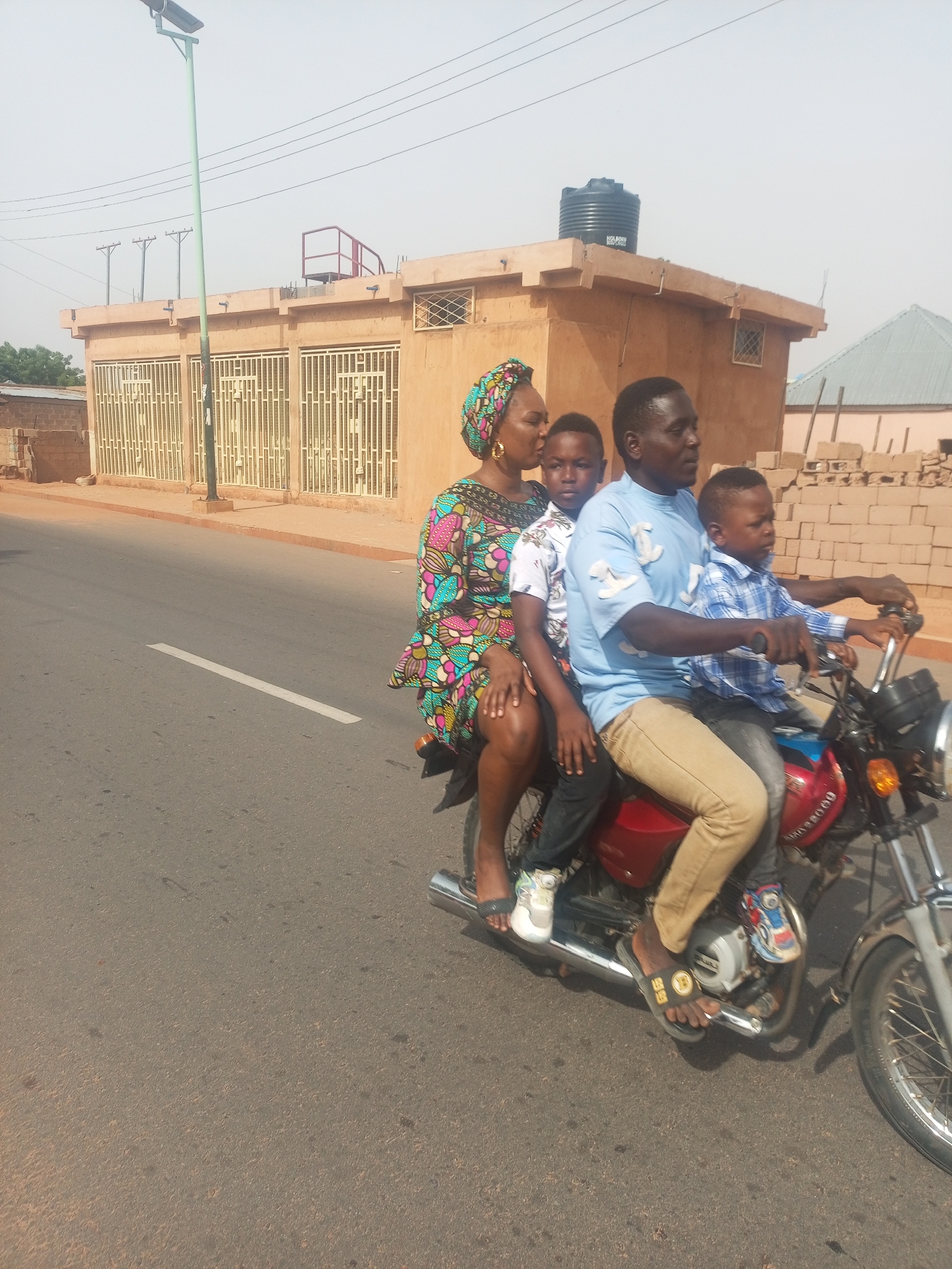 A man drives a red motorcycle on a street in Tempure, Nigeria. Seated behind him is a woman wearing a colorful dress and head wrap, and two young boys, all without helmets. The man wears a light blue t-shirt and tan pants, and sandals. The boy directly behind him wears a white shirt and black pants, and white sneakers. The other boy wears a blue plaid shirt and grey pants. The road is asphalt, with a white line dividing lanes. Buildings stand on either side of the road. The sky is hazy and bright. A water tank sits on the roof of a building in the background.