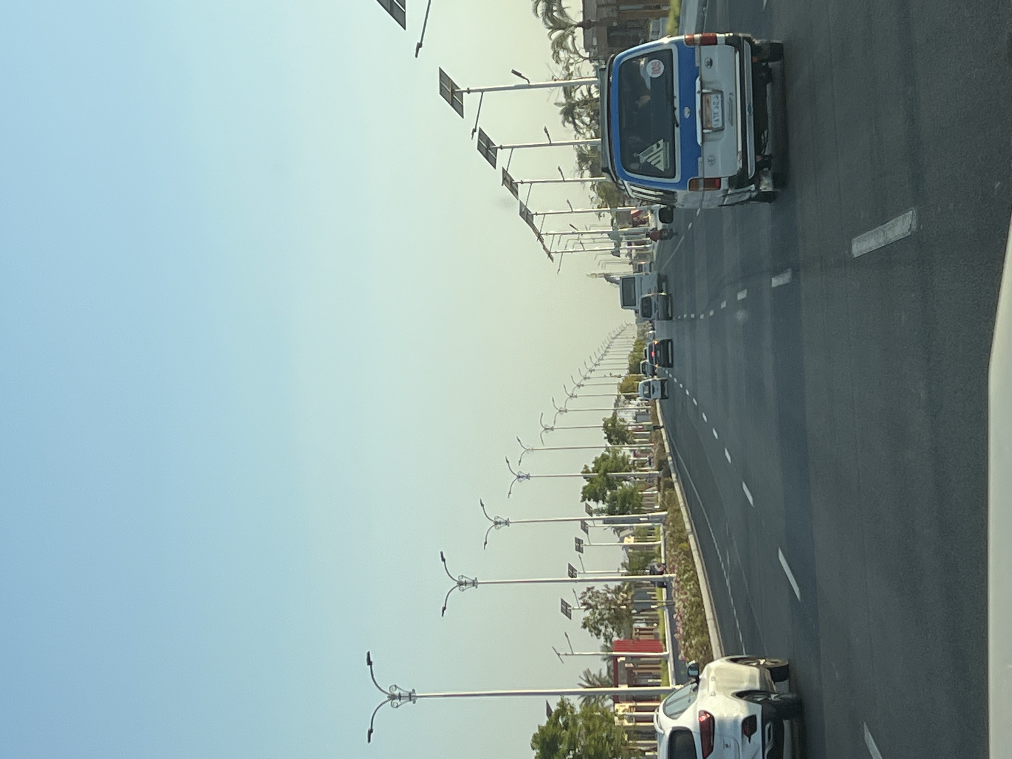 The image depicts a broad, paved road in Sharm El Sheikh, Egypt, lined with numerous modern streetlights featuring ornate designs and solar panels. The sky is a clear, pale blue, suggesting daytime with potentially warm weather. The road is multi-lane and appears to have distinct driving lanes demarcated by white lines. Several vehicles are present, including a white SUV in the left foreground, a blue and white van in the right foreground with visible license plates, a white bus further down the road, and other cars and motorcycles. On the sides of the road, there are manicured flower beds, green trees, and some palm trees, indicating a well-maintained urban environment. Buildings with red facades are visible in the background on the left, suggesting commercial or residential development. The overall scene conveys a sense of order and modernity in the Egyptian city.