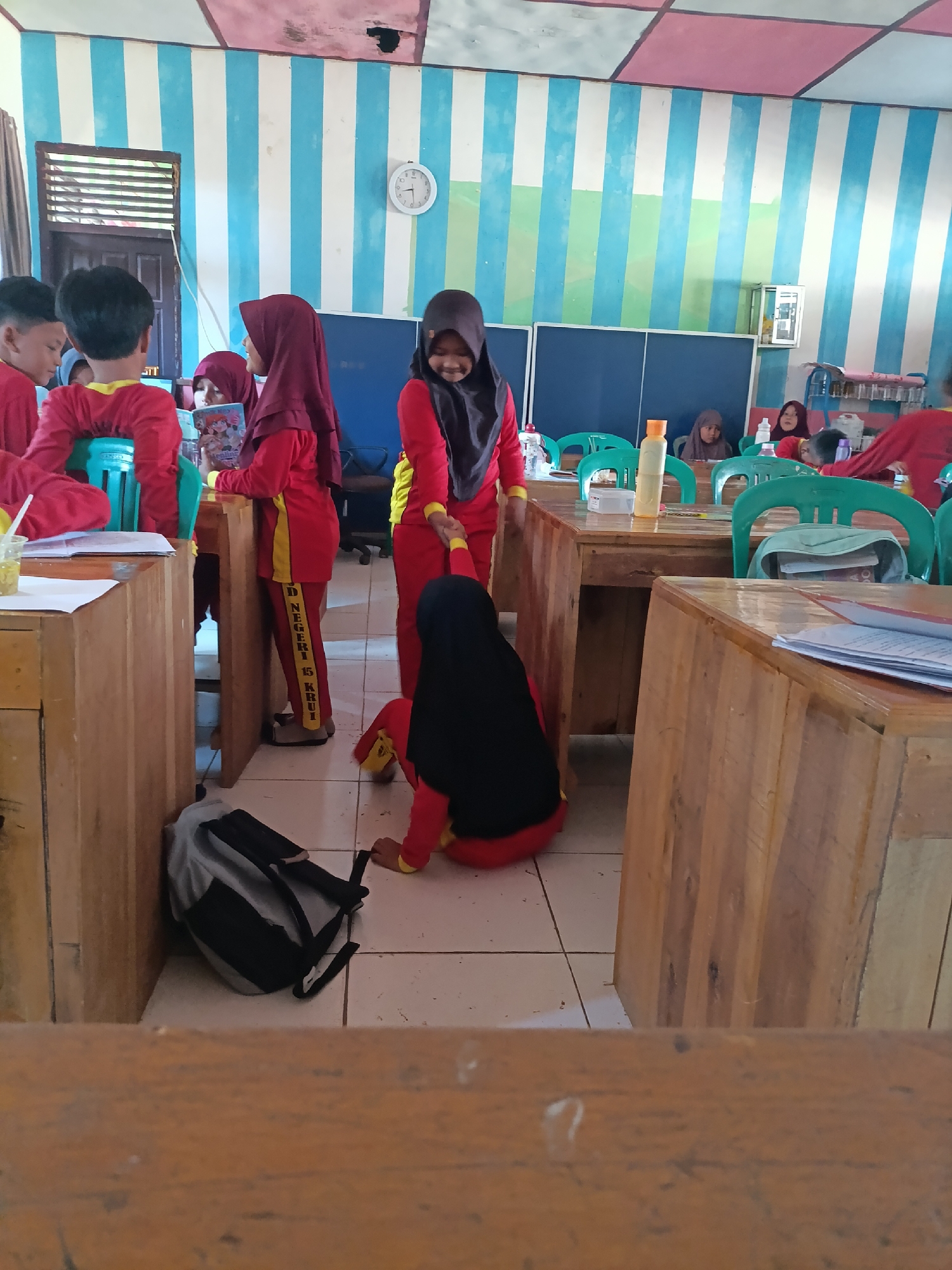 This image depicts a classroom scene in Sumberejo, Indonesia. Several young students, predominantly girls wearing red uniforms and hijabs, are visible. Some are seated at wooden desks, while others are standing. One girl in a burgundy hijab is holding a book and appears to be interacting with another student. A girl in a black hijab is kneeling on the floor. The classroom walls are decorated with vertical stripes in blue and white, and the ceiling has colored stripes. A clock is visible on the wall. Wooden desks and green plastic chairs are arranged in the room. A backpack is on the floor next to a desk. The overall atmosphere suggests a school environment during what appears to be a lesson or break.