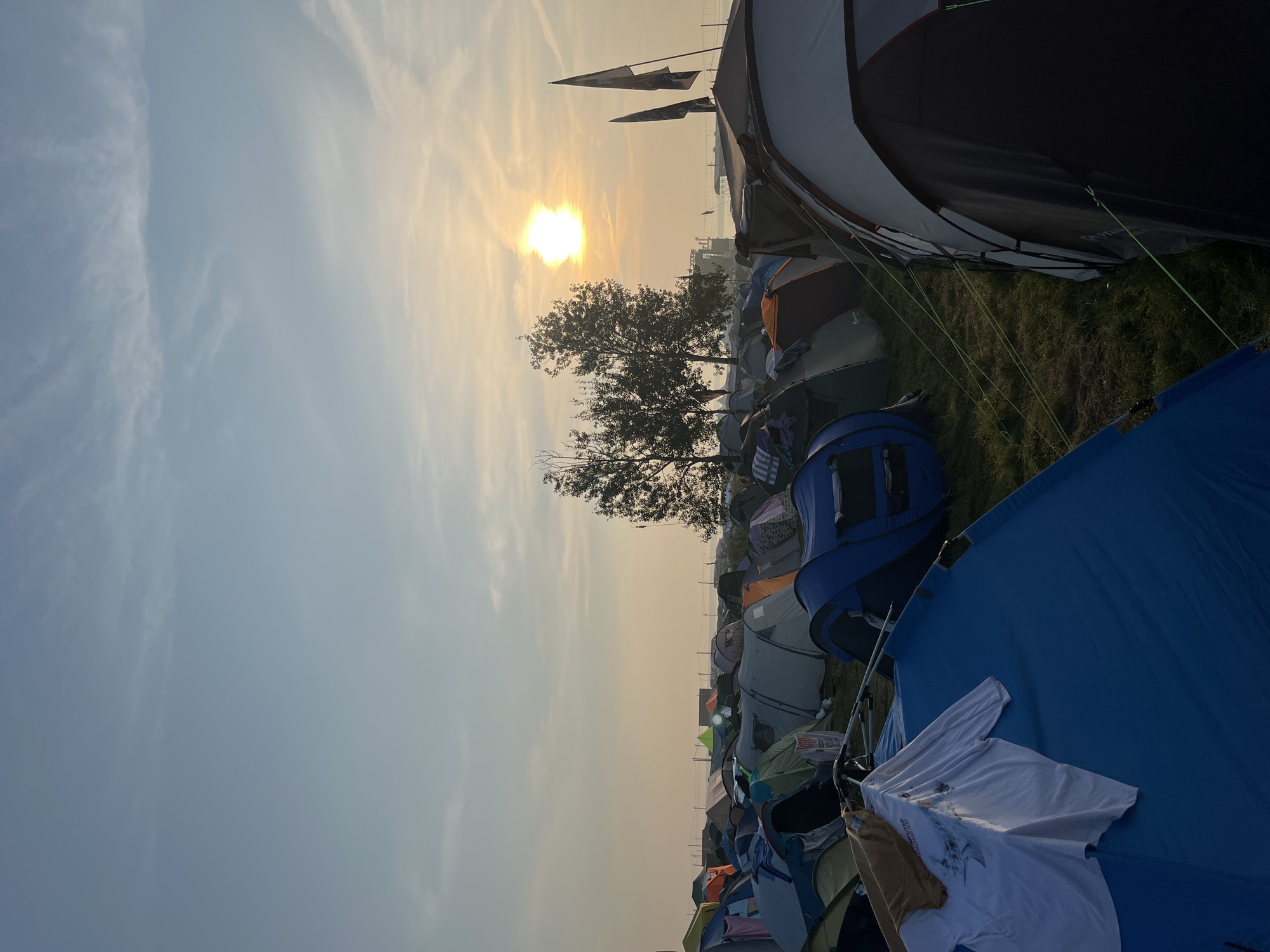 The image depicts a large campsite with numerous tents set up on a grassy field. The sun is rising or setting, casting a warm glow and creating a hazy atmosphere with some clouds in the sky. There are several trees in the mid-ground, and in the distance, there are some structures and possibly a body of water. The foreground shows a close-up of several tents, with some clothes hanging on a tent pole in the lower left. The scene appears to be a temporary setup, likely for an event or festival, given the density and variety of the tents. The location is identified as Surakovo, Russia.
