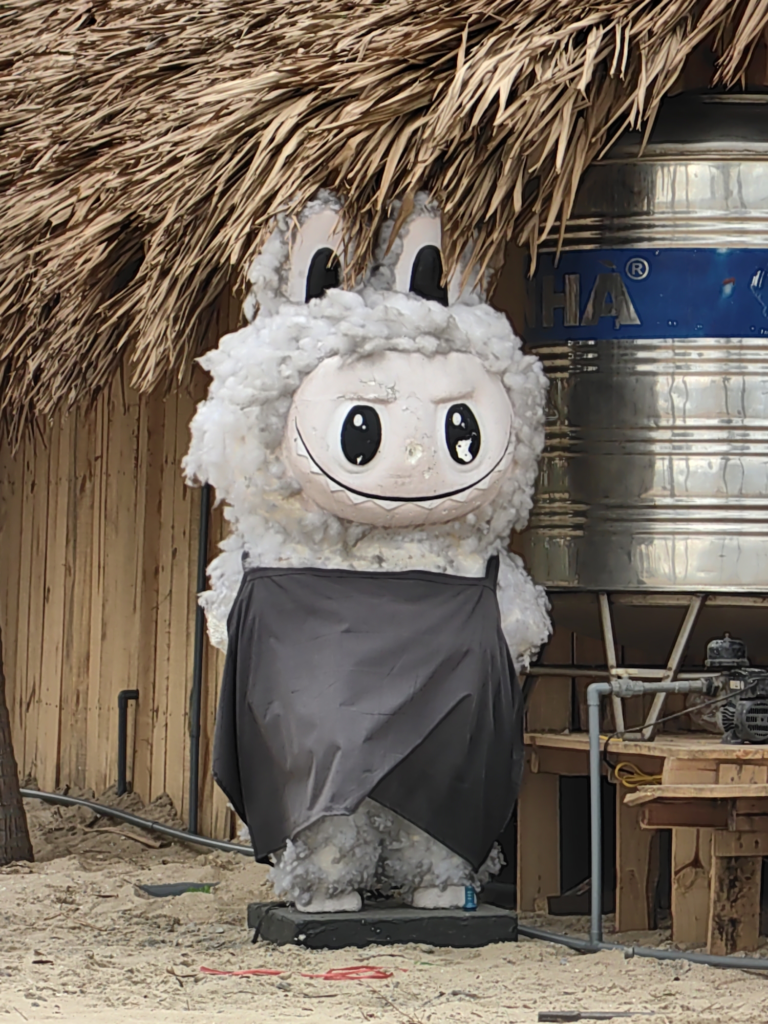 The image shows a large, cartoonish mascot figure, resembling a fluffy sheep or lamb, wearing a black apron. The figure is situated outdoors, likely in a sandy area, possibly a beach. Behind the mascot is a wooden structure with a thatched roof, reminiscent of a beach hut or a small building for a resort. To the right of the figure, a large, cylindrical stainless steel tank is visible, with blue lettering that includes the letters "HA". Pipes and what appears to be a small pump or motor are connected to the tank. The overall scene appears to be set in a coastal or tropical environment, consistent with a location in Bai Chay Ward, Vietnam.