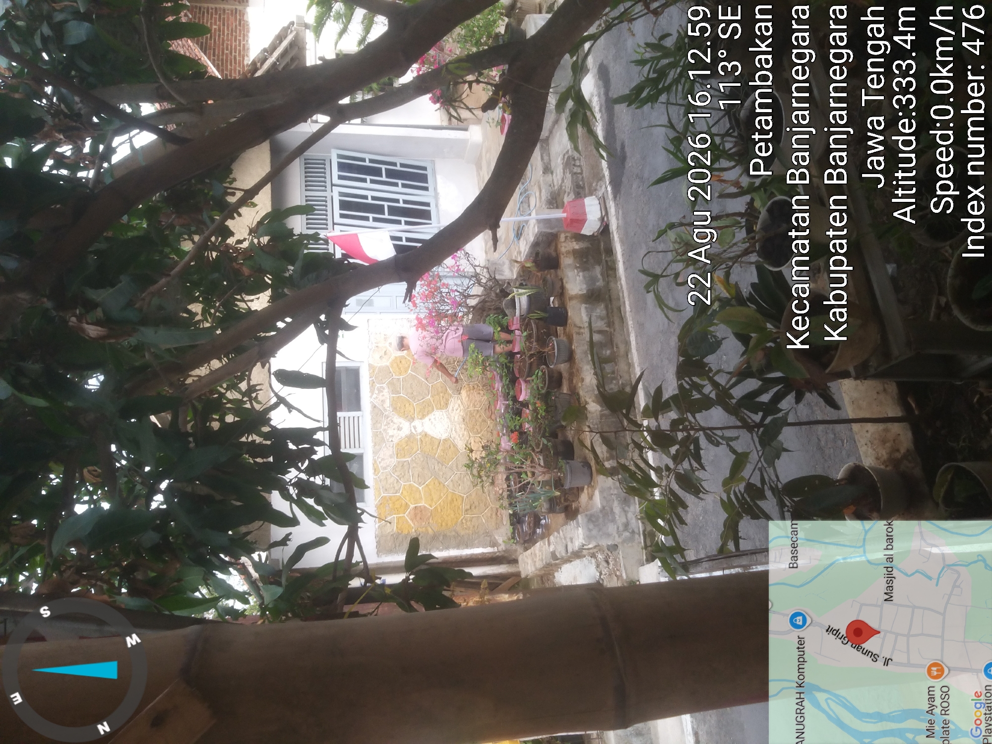 The image displays a scene in Petambakan, Banjarnegara Regency, Central Java, Indonesia. A person in a pink shirt and shorts is watering plants in a garden. The garden features numerous potted plants, some in plastic containers and others in terracotta pots. The house in the background has a distinctive textured wall with a stylized image and a red and white Indonesian flag displayed on the wall. A small map inset in the lower left corner shows a location marked on Jl. Sunan Gripit, indicating a specific area within Petambakan. Overlaid on the image are timestamps, coordinates, and other data, including the date "22 Agu 2026 16.12.59", an altitude of "333.4m", and a speed of "0.0km/h". The overall environment suggests a residential outdoor setting with lush greenery.