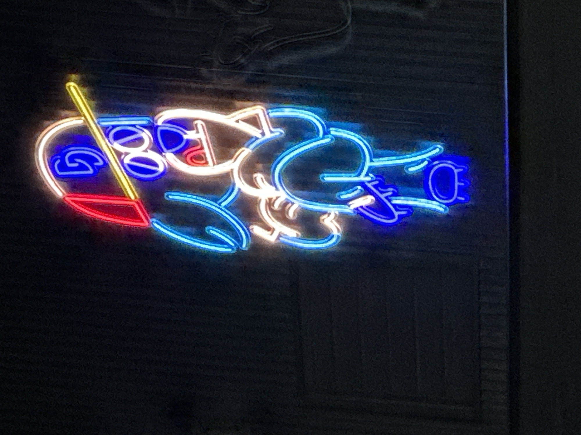 A nighttime shot shows a neon sign depicting what appears to be a cartoon character, possibly an alien or a creature with exaggerated features, illuminated with neon tubing of various colors, including blue, red, yellow, and white. The sign is attached to a dark wall, possibly made of dark-colored planks. The location is given as Unknown, Unknown. The specific details of the character's design or the exact nature of the scene the sign is intended to portray are not apparent from the image.