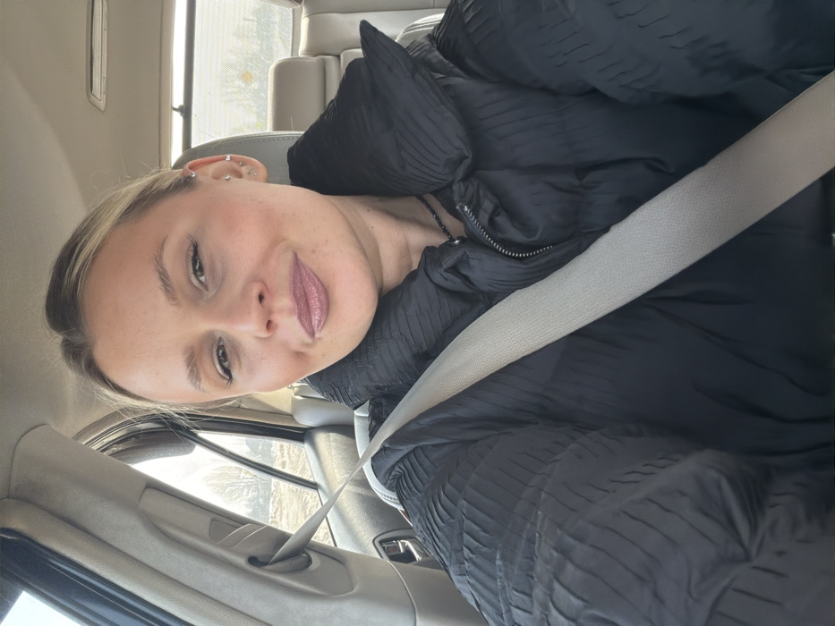 An adult female, appearing to have light blonde hair pulled back, is depicted in a close-up selfie taken from the passenger seat of a vehicle. She wears a black puffer jacket with a textured, pleated design and a light-colored seatbelt extending diagonally from her left shoulder across her chest. A dark beaded necklace is visible around her neck. Her left ear displays multiple piercings. Her facial expression is a slight smile, with visible makeup including defined eyebrows, mascara, and medium-toned lipstick. The vehicle's interior features light-colored upholstery and door panels. Through the car's passenger window, an exterior scene of an undeveloped area is visible, characterized by bare trees and exposed ground under natural daylight. This scene is located in the city of Дулесово, Russia.