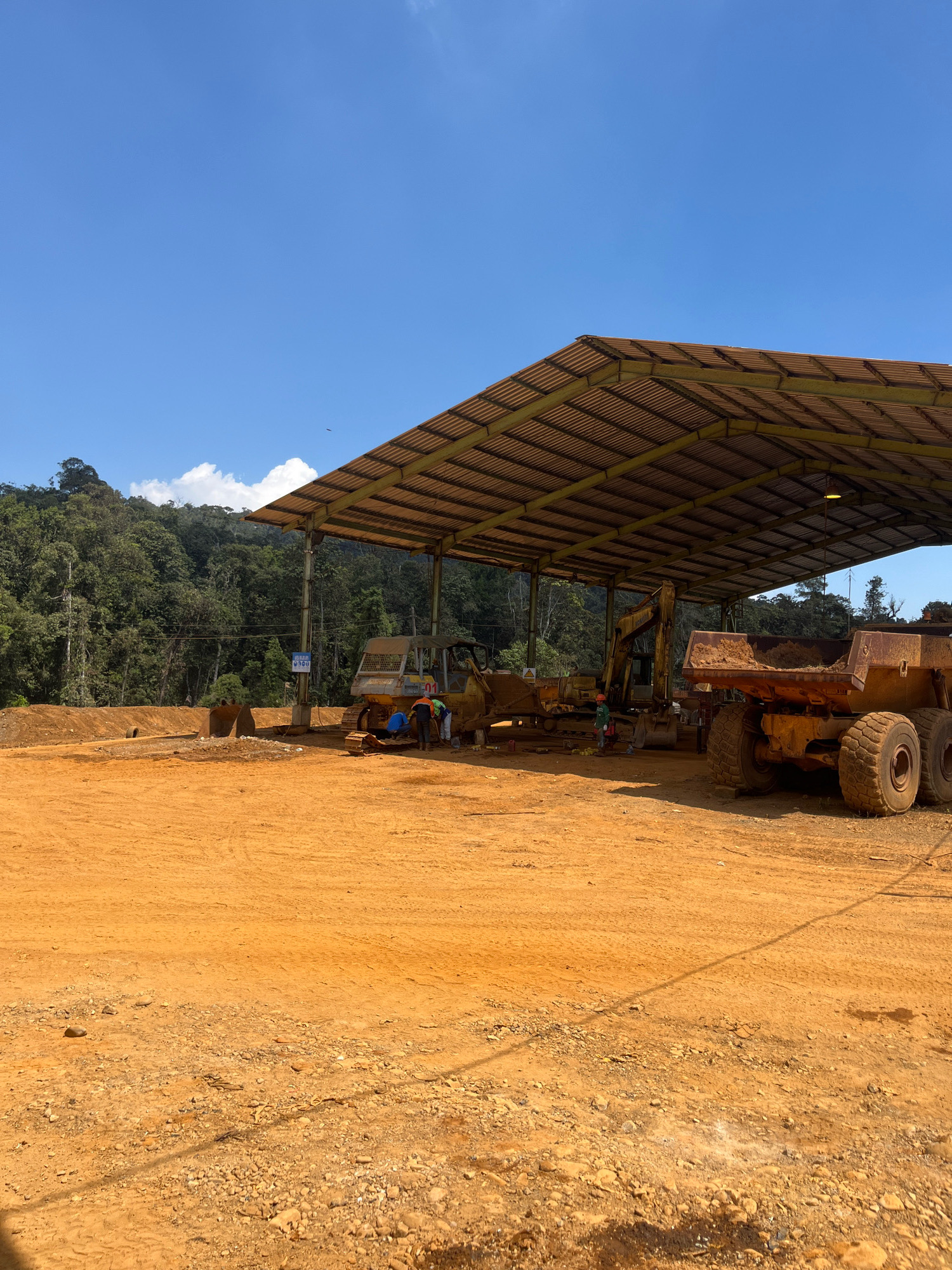 A wide shot captures an outdoor area at a construction or mining site in an unknown location in Indonesia. The foreground is dominated by orange-brown dirt, with some gravel and scattered rocks. In the midground, a large, covered structure with a corrugated metal roof supported by metal poles provides shade. Underneath this structure, several heavy machinery vehicles are parked, including a bulldozer and a large dump truck filled with dirt. A few individuals, appearing to be workers, are visible near the machinery, some of whom seem to be engaged in repair or maintenance. A blue sign with white text is mounted on a pole near the machinery. In the background, a lush, green, densely forested hillside rises under a bright blue sky with scattered white clouds. The overall impression is that of an active, but perhaps paused, work area within a tropical environment.