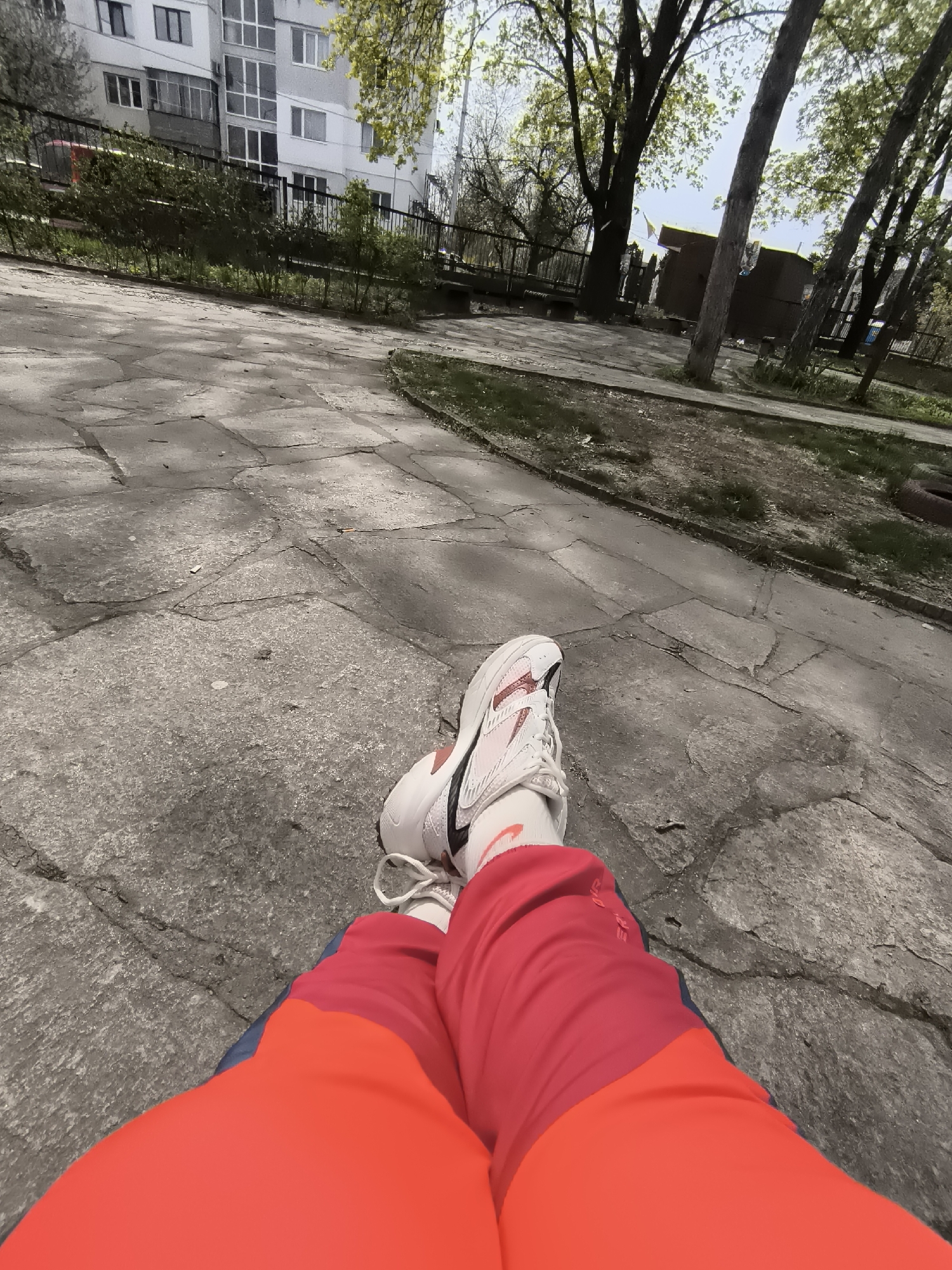 A first-person perspective image shows someone sitting on a concrete surface in an urban park. The individual is wearing a pair of white Nike sneakers with a red swoosh, white socks with a red Nike swoosh, and bright red and blue athletic pants. The concrete is cracked and uneven.
In the background, there is a gray apartment building with multiple windows and balconies. A black metal fence runs along the edge of a grassy area with trees and bushes. The trees are partly bare, suggesting either early spring or late autumn. Other buildings are visible in the far background, and there is a hint of sky. The photograph was taken in the city of Pleven, Bulgaria.