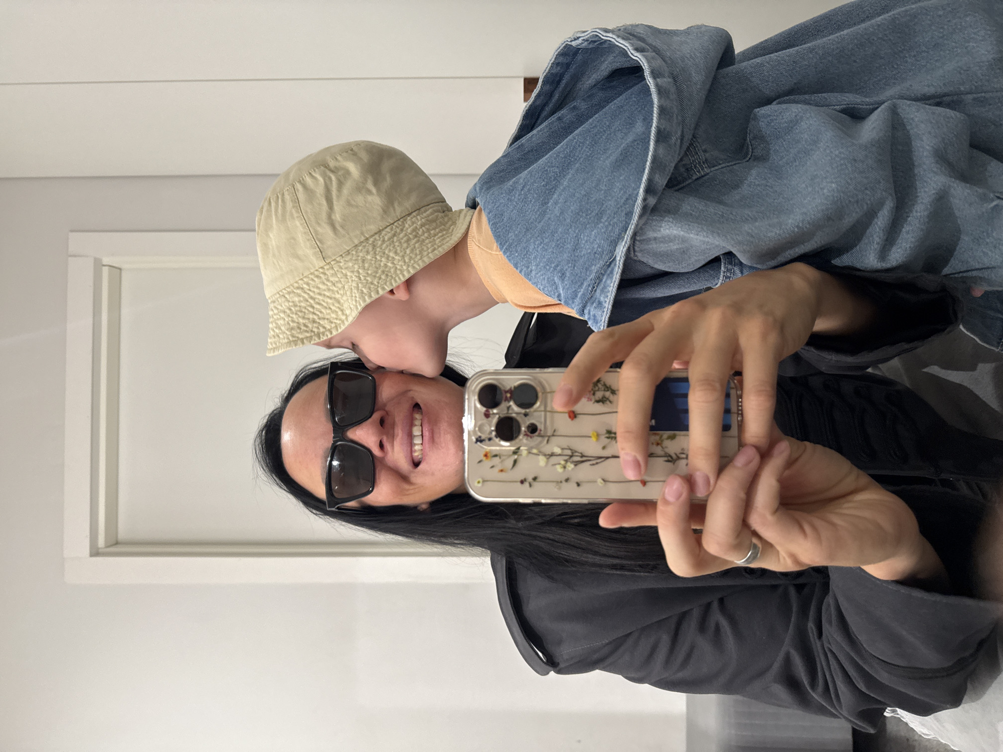 In a dimly lit indoor setting, a person wearing sunglasses and a black jacket takes a selfie. A child in a light-colored bucket hat and denim jacket kisses the person's cheek. The person is holding a smartphone with a floral-patterned case. The background appears to be a wall with some molding.