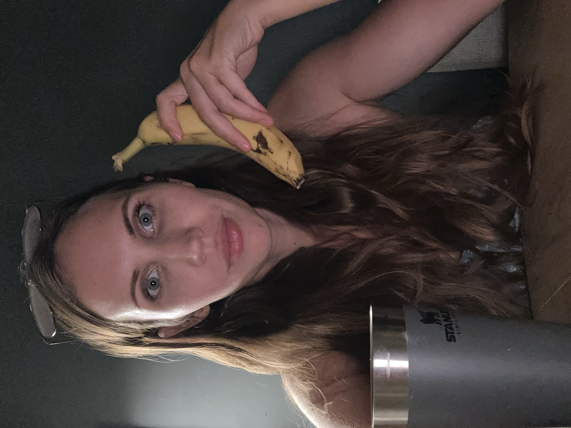 A medium shot shows a woman holding a banana to her ear like a phone. She is positioned in the center of the frame, with her face filling most of the image. She has long, wavy brown hair that falls around her shoulders. Her eyes are wide and blue, and she is looking directly at the camera. She is wearing a grey tumbler with the Stanley logo in front of her. The background is dark and appears to be an indoor setting, possibly a dining room or office. There is no indication of Fethiye, Turkey in the image.