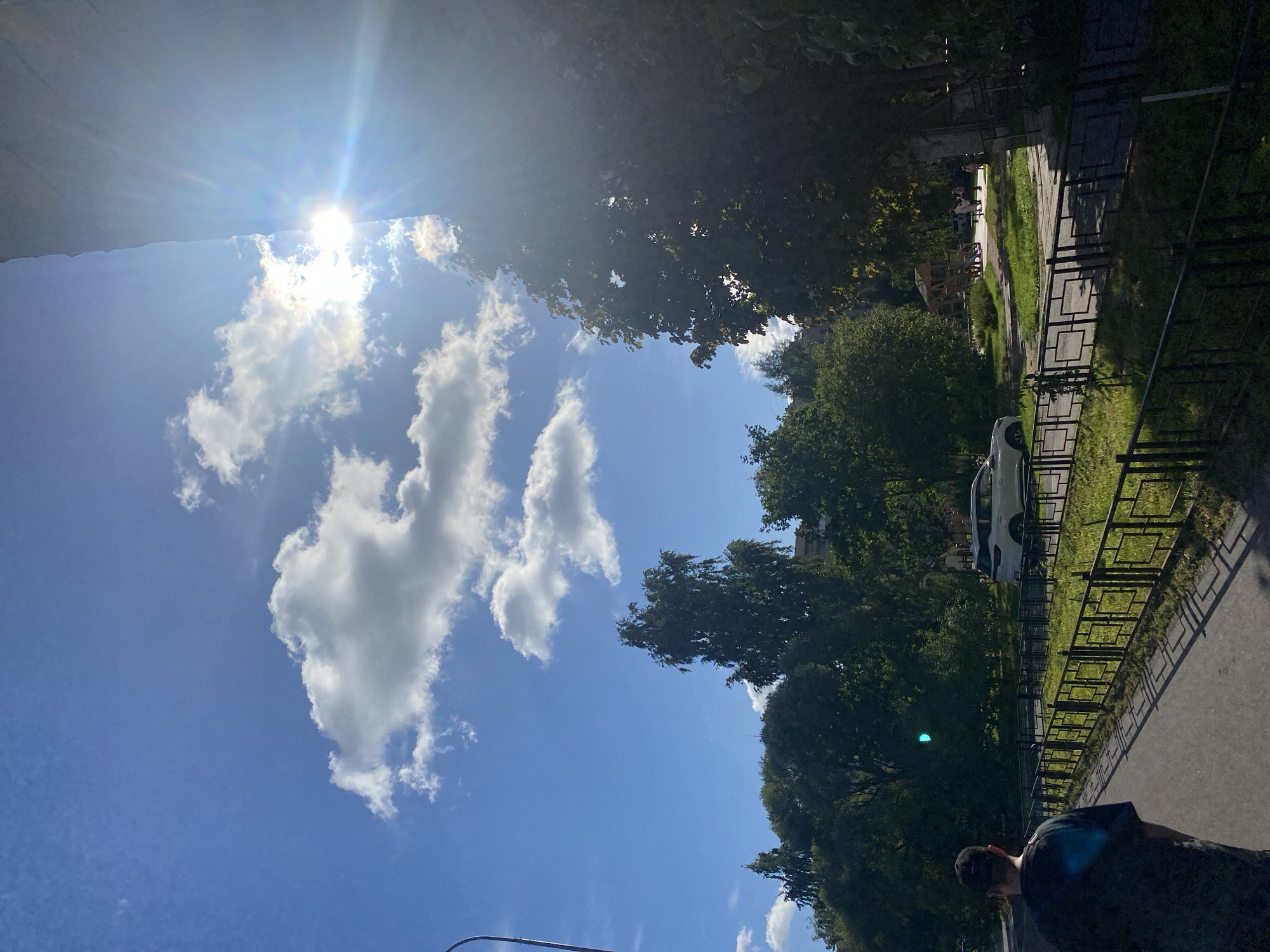 In Saint Petersburg, Russia, on a sunny day, a person is walking down a sidewalk next to a black metal fence. Beyond the fence, there is a green lawn, a road with a white car, and a playground with children. The sun is shining brightly, casting long shadows. Tall trees line the street, and there are buildings in the background. The sky is blue with scattered white clouds.