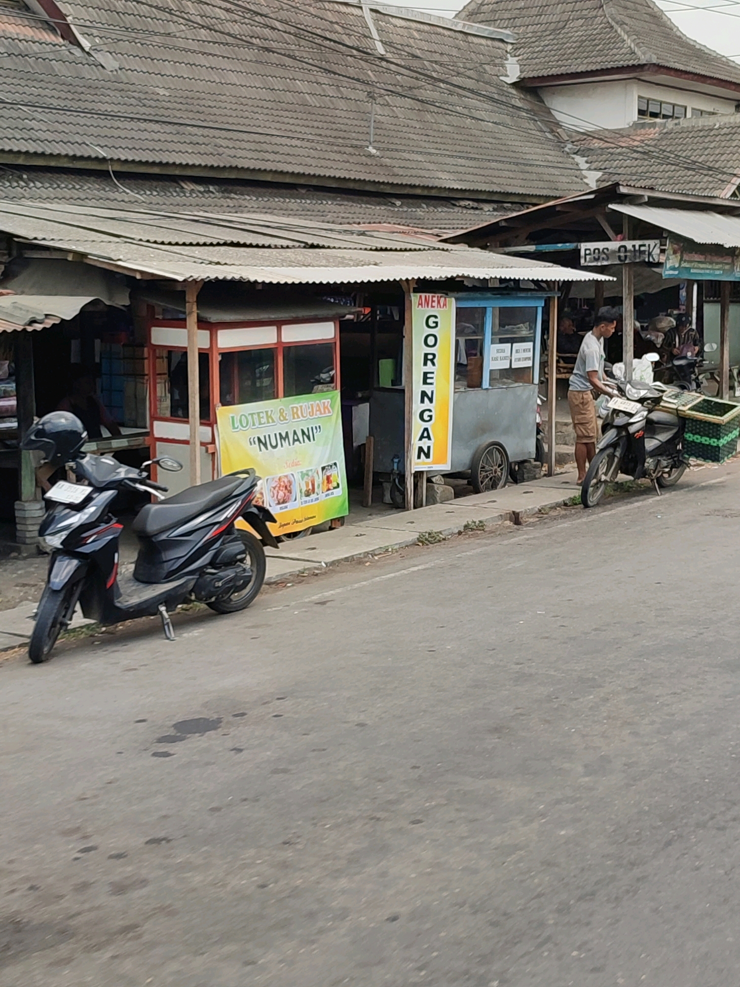 This is a photograph taken in Salaman, Indonesia. The scene depicts a street with several food stalls and parked motorcycles.
In the foreground, a black scooter is parked on the right side of the road. It has a license plate that appears to be Indonesian. A black helmet rests on the handlebars. Further down the road, another motorcycle is parked with a large green basket attached to the rear. A person in a light-colored t-shirt and tan shorts is standing next to this motorcycle, appearing to interact with a food stall.
The food stalls are set up along a sidewalk. One stall has a yellow banner with Indonesian text, including "ANEKA GORENGAN" and "LOTEX & RUJAK 'NUMANI'". This stall appears to be selling fried foods and rujak. Next to it is another stall with a blue and grey cart. In the background, a building with a tiled roof is visible. The street itself is paved and appears to be moderately used. The environment suggests a typical street scene in a Indonesian town, likely a commercial or market area.