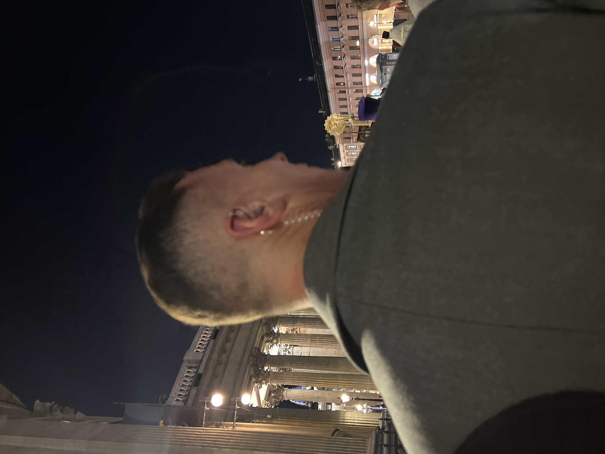 A medium shot shows a man in the foreground, facing away from the camera. He has a short haircut, and part of his face and neck are visible. He wears a light-colored necklace. He is wearing a dark gray jacket.
Behind him, the image includes illuminated historical buildings with columns, and a distant building with multiple windows. The sky above is dark, suggesting it is night. The architectural details suggest a location in the city of Saint Petersburg, Russia, specifically the Winter Palace and Palace Square. There is evidence of street lighting.
