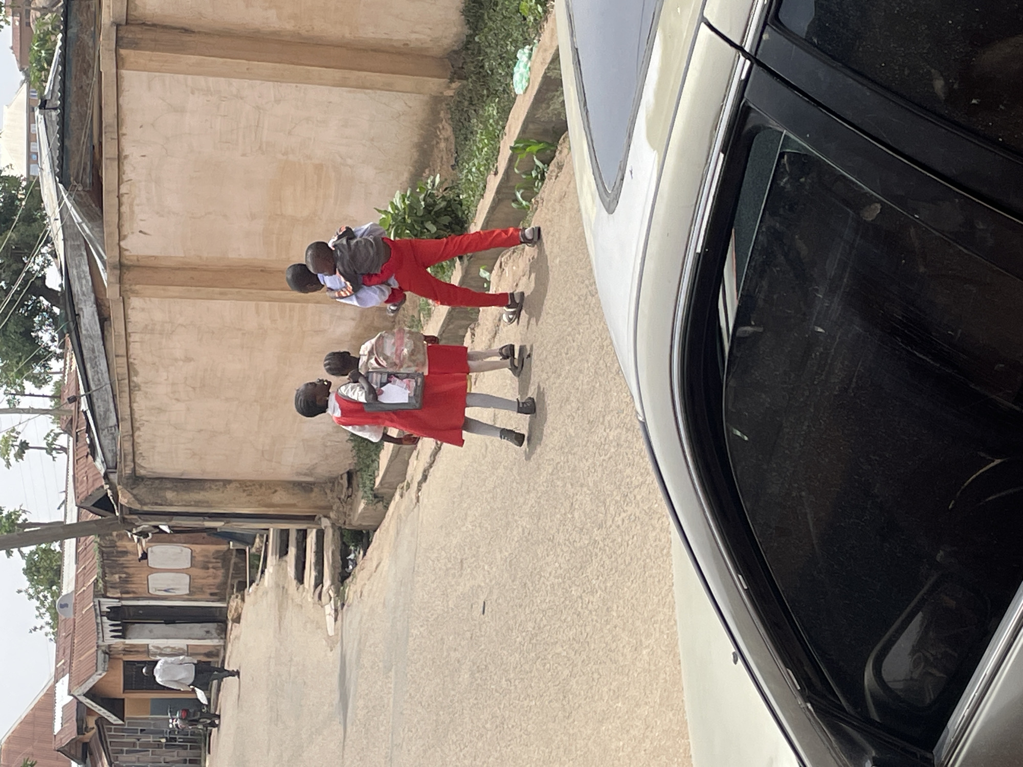 This image, rotated to its correct vertical orientation, captures a daytime street scene in what appears to be a residential or semi-urban area. The sun is bright, suggesting clear weather.
The primary subjects are a group of young people, likely school children, walking along a road or path. In the foreground, slightly to the left, are two girls. Both wear what appears to be a school uniform consisting of a white shirt, a red dress or skirt, and dark shoes, and each carries a backpack. Behind them, a boy in red trousers and a light-colored shirt, also with a backpack, is giving another child a piggyback ride. The child on his back wears a light-colored shirt.
To the right, a white car occupies a significant portion of the frame, showing its side and a dirty window. The car appears parked or moving slowly alongside the road. The road surface itself is light-colored and seems to be a mix of dirt and possibly gravel or a rough paved surface.
On the left side of the road, a narrow concrete drainage ditch runs parallel to the path, with some green vegetation growing along its edge. Beyond the ditch are various buildings and structures. These buildings feature concrete walls, some with visible plaster, and several have corrugated iron roofs. Further down the street, a person in a white shirt is visible, standing near a motorcycle or scooter. The overall environment suggests a bustling community setting.
No discernible text is visible on any of the subjects, uniforms, vehicles, or buildings.