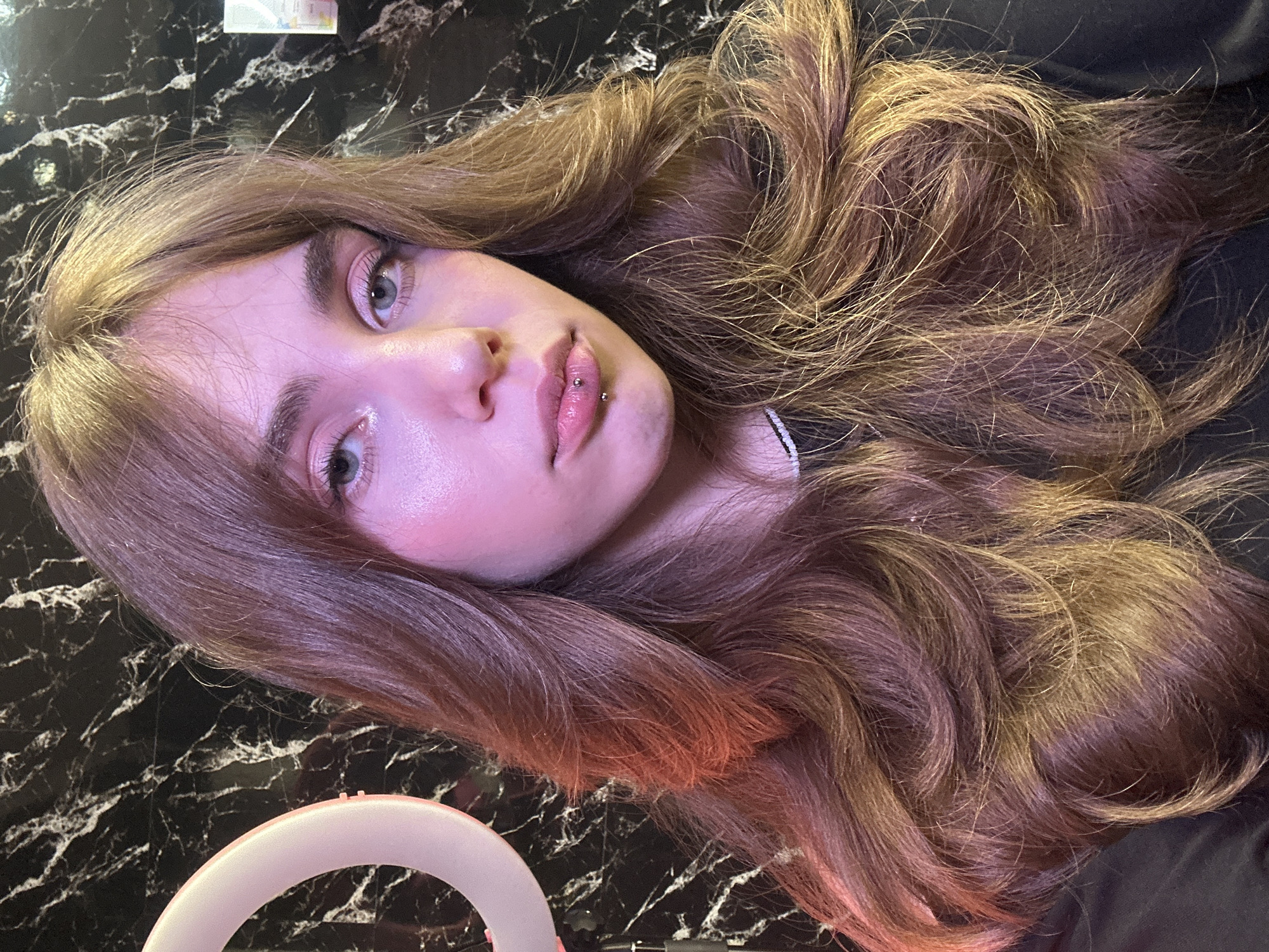 A young woman with long, wavy brown hair and blue eyes is pictured from the chest up. She has light skin with pink blush on her cheeks. Her lips are slightly parted, revealing a monroe piercing. She is wearing a black shirt. The background is a black and white marbled pattern. To the left of the woman's head, a pink ring light is visible. The image appears to be taken indoors, likely in a home setting, given the ring light and backdrop, which are common for content creation. There is no specific indication of the city of Saratov, Russia.