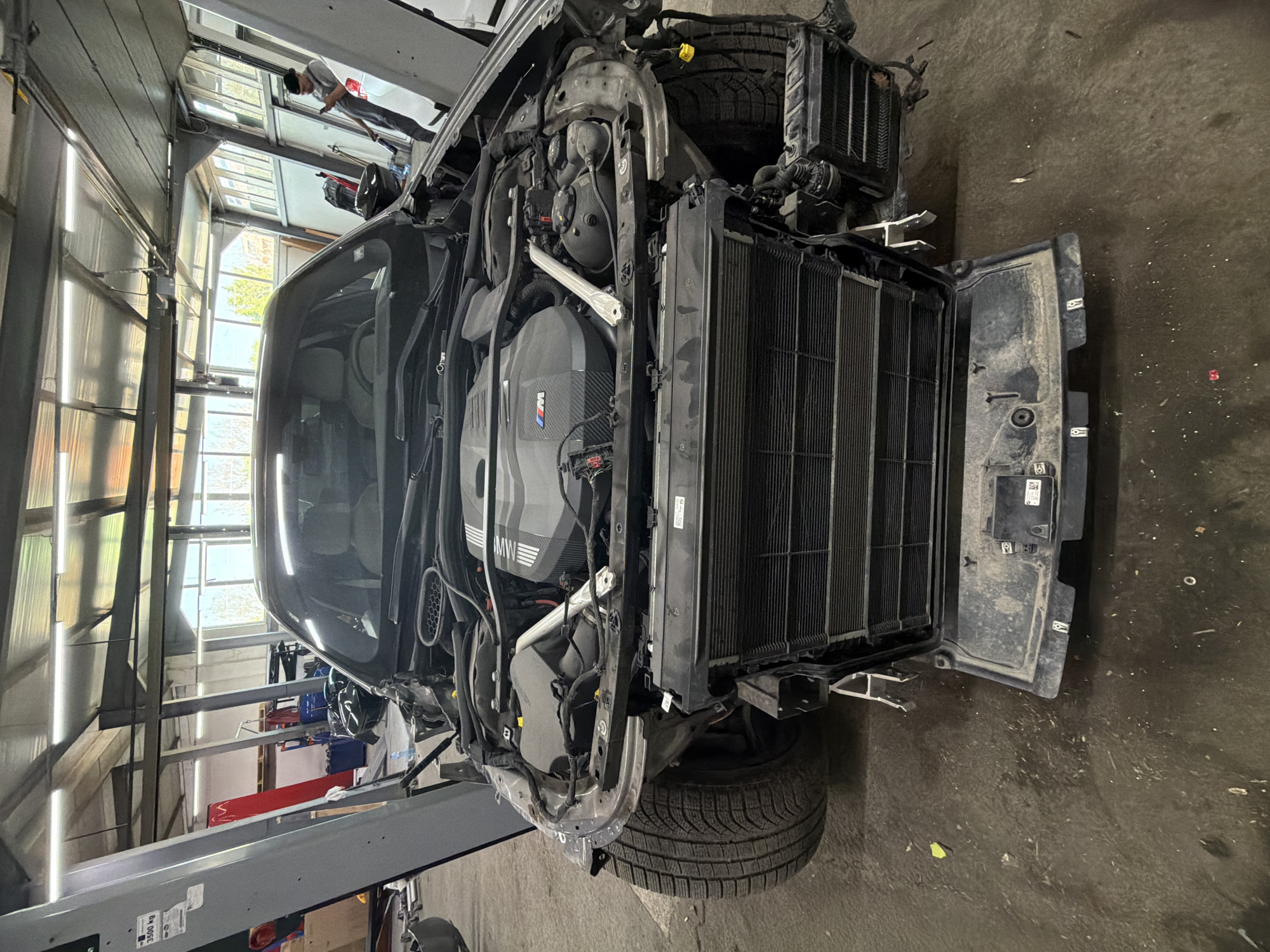 The image shows the front of a modern car, possibly a BMW M series, with its front body panels and bumper removed, exposing the engine, radiator, and other mechanical components. The car is elevated on a lift inside a repair shop or garage, with various tools and equipment visible in the background. The setting appears to be in a workshop or a mechanic garage, with industrial lighting and a concrete floor. There is no specific information to confirm it is taken in Burgas, Bulgaria. No people are clearly visible in the immediate foreground.