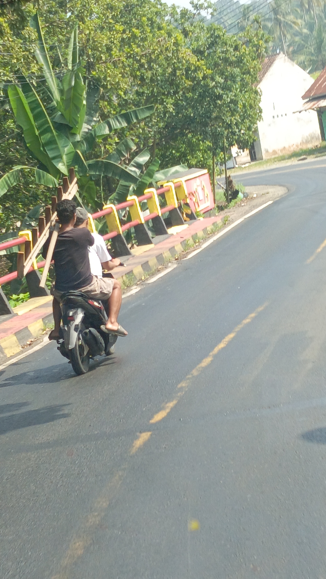 Two individuals are riding on a motorcycle on a paved road. The person in the back is wearing a dark t-shirt and light-colored shorts, and the person in front is wearing a white t-shirt and a light-colored cap. They are traveling on a road that curves to the right. On the left side of the road, there is a yellow and red guardrail with a black and yellow striped base. Behind the guardrail, there are lush green trees and banana plants. In the background, a white building with a red-tiled roof is visible, situated on a slight incline. The scene appears to be in a rural or semi-rural area of Kota Agung, Indonesia, given the vegetation and building style. The road has a yellow dashed line painted down the center. The lighting suggests it is daytime, with shadows cast on the road.