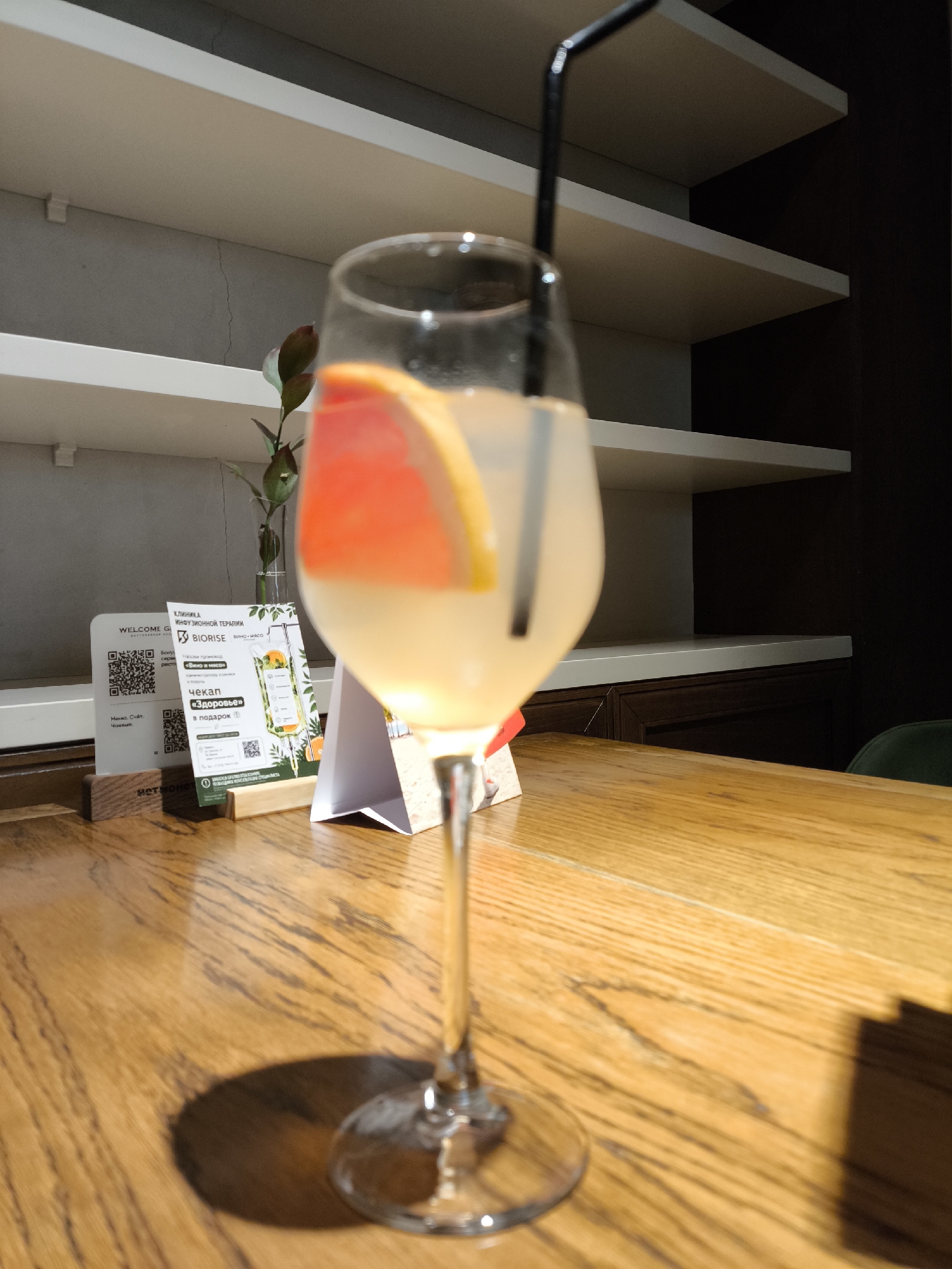 An indoor scene in a restaurant or cafe in Izhevsk, Russia, features a stemmed glass containing a light yellow beverage with a black plastic straw and a large slice of red grapefruit, positioned slightly right of center on a light-colored wooden table. The glass casts a prominent shadow to its left. On the table to the left, a clear vase holds a small plant with dark green leaves. Adjacent to the plant are two informational stands: a white stand with a "WELCOME G" header and QR codes, and a green and white flyer advertising "BIORISE Clinic of Infusion Therapy" with "WINE + MEAT" and a "health check-up as a gift" promotion, confirming the Izhevsk location. The background consists of light grey walls with multiple white floating shelves. A distinct vertical crack is visible on the grey wall on the left. The right background features a dark brown cabinet or wall beneath the lower shelves, and a portion of a dark green upholstered chair is visible on the far right. No individuals are present.
