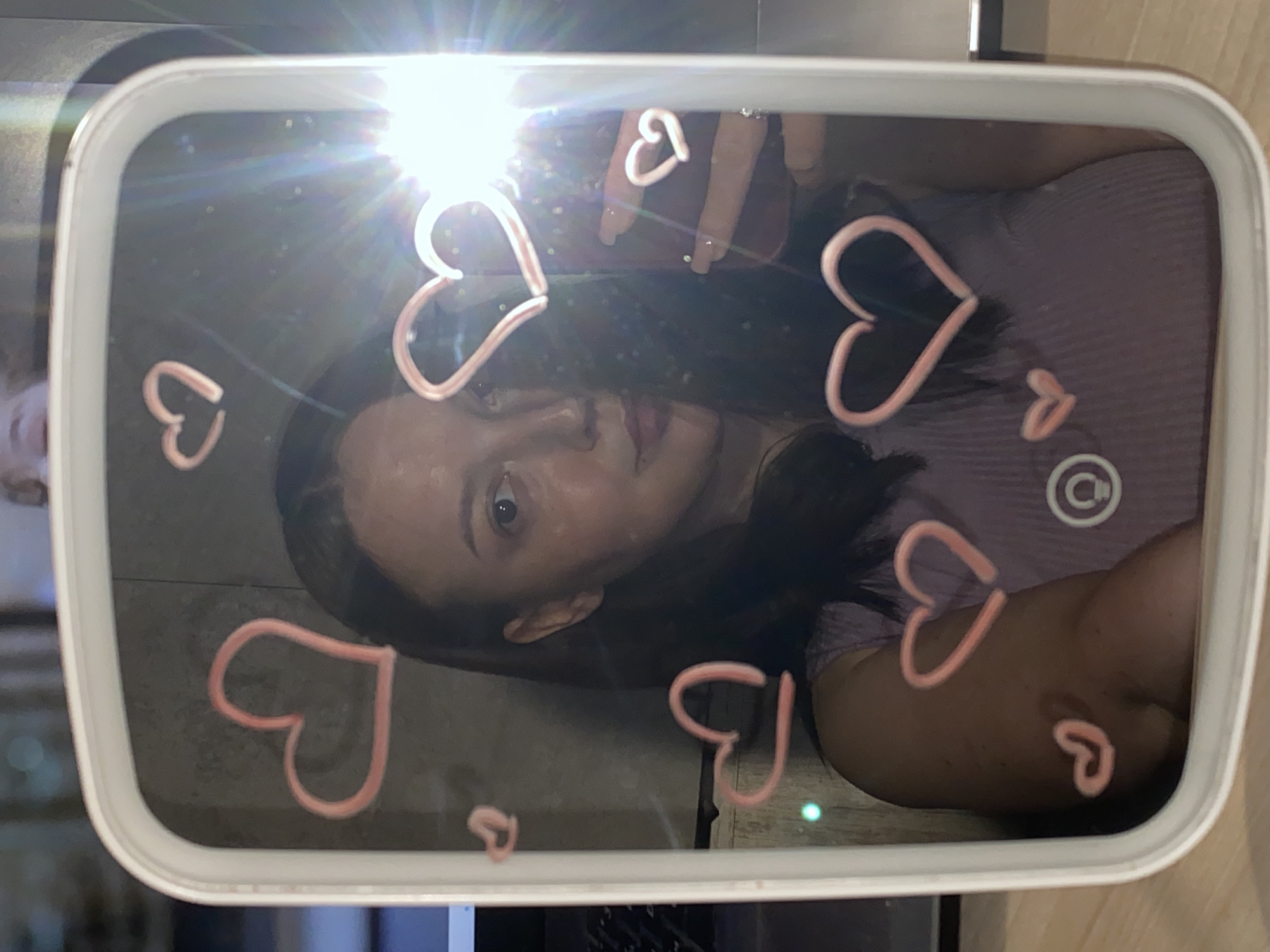 A woman is taking a selfie in a mirror decorated with pink hearts. The location is unknown, and there is no indication of Chaikovsky, Russia. The woman is holding a phone with her right hand, which can be seen at the top of the frame. She is wearing a purple shirt and has dark hair. The mirror is white and reflects a portion of the room. The mirror has a white frame.