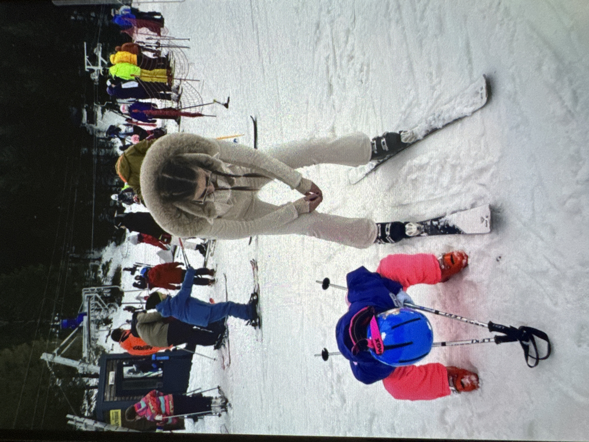 The image depicts a snowy ski slope at a recreational area in Vratsa, Bulgaria, during daytime. In the foreground, a young child wearing a blue jacket, bright pink pants, and red ski boots is crouched on the snow, with a blue helmet featuring a pink accent on their head. Two ski poles are planted upright in the snow directly in front of the child. Standing over the child, leaning forward, is an adult female dressed in a light beige/cream-colored fur-hooded ski suit, dark ski boots, and skis on her feet. Her hands are clasped in front of her, and she wears glasses with her hair in braids.
In the midground, multiple individuals in various colored ski apparel (including orange, olive green, blue, yellow, red, and purple jackets) are visible standing on skis, suggesting a gathering area or queue for a lift. Ski poles and skis are associated with these individuals. A red and white net or fence delineates a portion of the snowy area to the right.
The background features a prominent ski lift structure, specifically a chairlift tower, against a backdrop of dark, dense evergreen trees. A small building, possibly a lift station or service hut, is located to the left of the main lift tower. The entire ground is covered in snow, showing subtle textures and tracks from ski activity. The overall lighting suggests an overcast day.