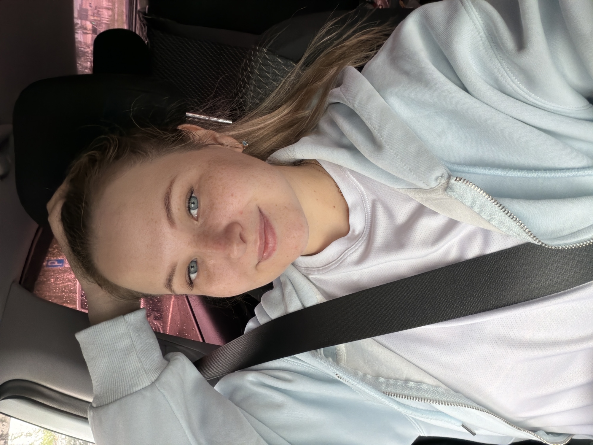 A fair-skinned young woman with freckles and light brown hair sits in the passenger seat of a car. She is wearing a white shirt under a light blue zip-up hoodie and a black seatbelt. Her left hand is placed on her head. The car appears to be stopped or moving slowly, as can be discerned from the raindrops on the passenger-side window. Outside the window, a blurry cityscape is visible, potentially representing Chaikovsky, Russia. The interior of the car, including the black headrest of the driver's seat, is visible. The scene depicts a casual, everyday situation.