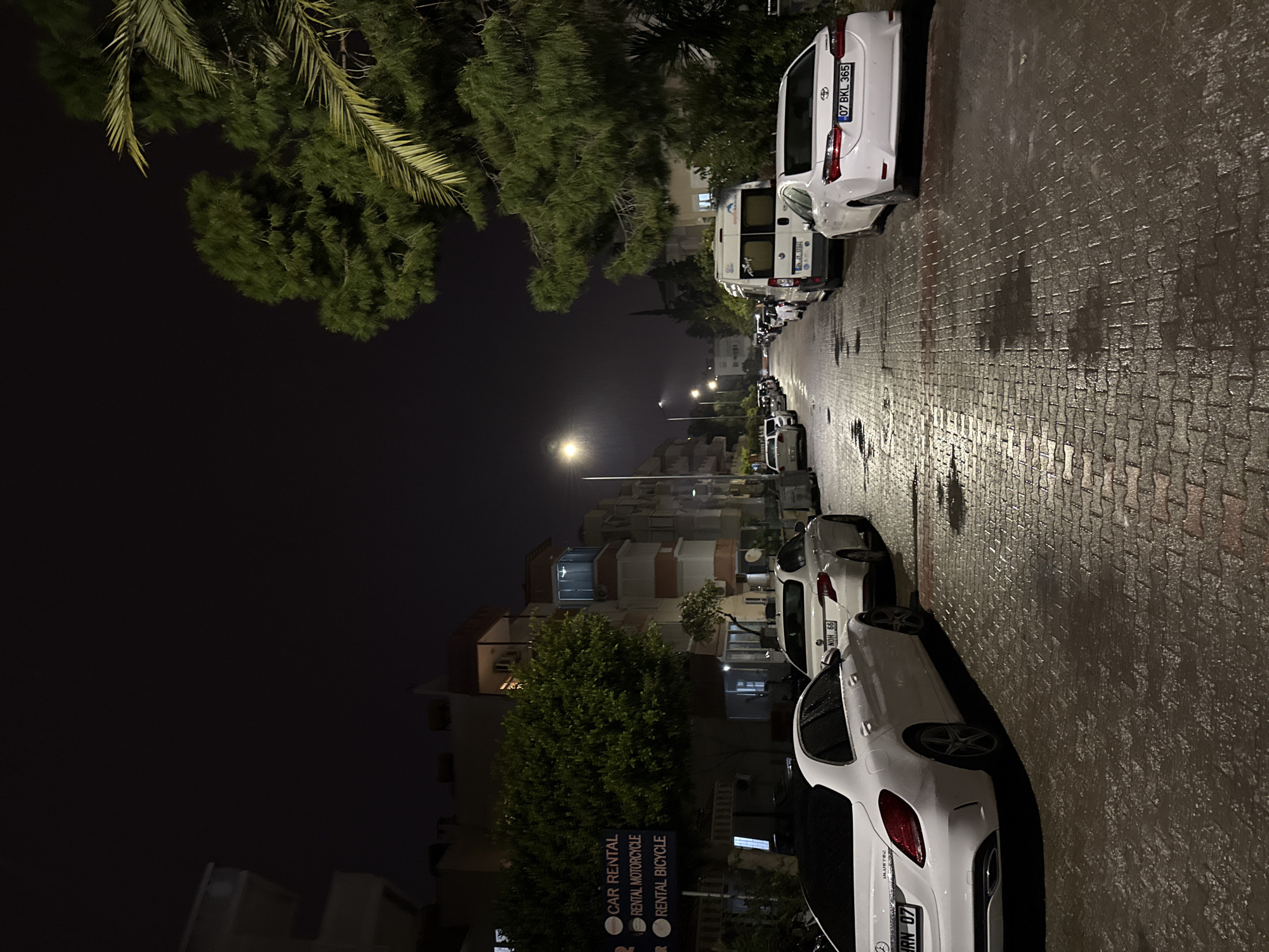 The image captures a night scene in Alanya, Turkey. A street lined with cars is visible, with a white Toyota Corolla parked on the right. Additional cars are parked along the street, including a white van and a Mercedes-Benz. Street lights illuminate the wet, brick-paved street, reflecting the artificial light. Buildings and trees line the street, and a sign for "CAR RENTAL, RENTAL MOTORCYCLE, RENTAL BICYCLE" is visible.