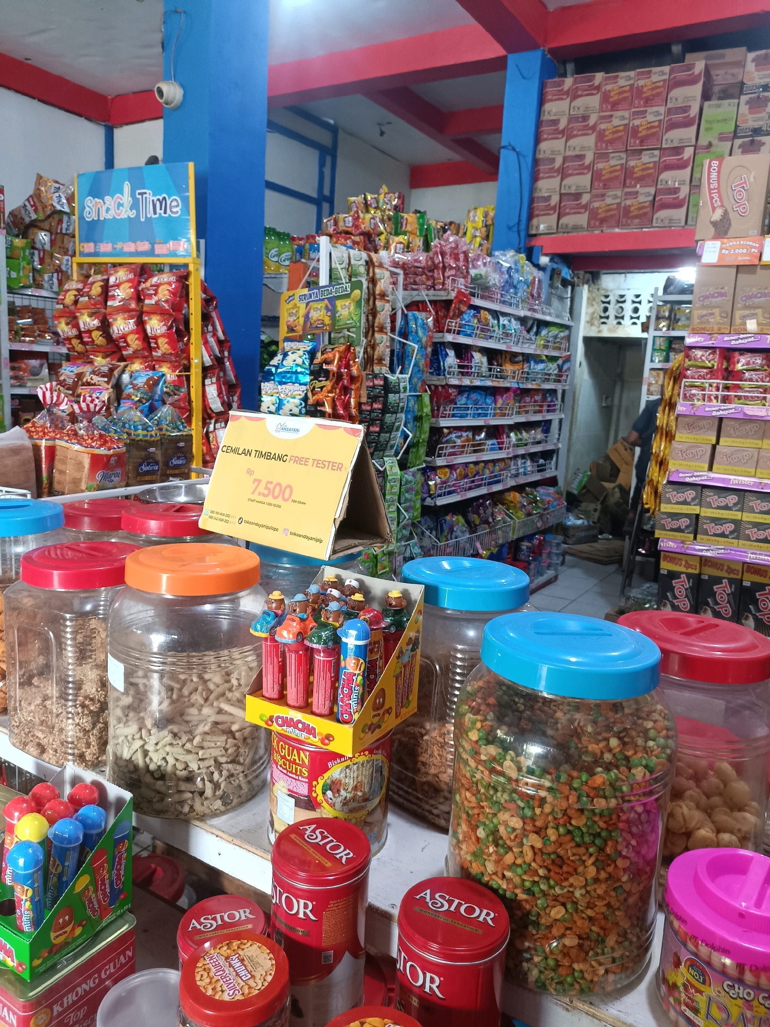The image depicts the interior of a small convenience store, likely a minimarket, in Palopo, Indonesia. The store is well-stocked with various packaged snacks and food items, displayed on shelves and in large clear plastic jars. Prominently featured in the foreground are several large clear jars filled with colorful snacks. To the left, a yellow sign with blue text reads "Snack Time" above shelves laden with bagged snacks. In the center, a yellow triangular display sign advertises "CEMILAN TIMBANG FREE TESTER" with a price of "Rp 7.500". Various red and blue lidded jars and containers of snacks are also visible. In the background, shelves are filled with more packaged goods, and a person is partially visible in the back of the store. The overall impression is of a brightly lit, organized retail space typical of a local Indonesian convenience store.