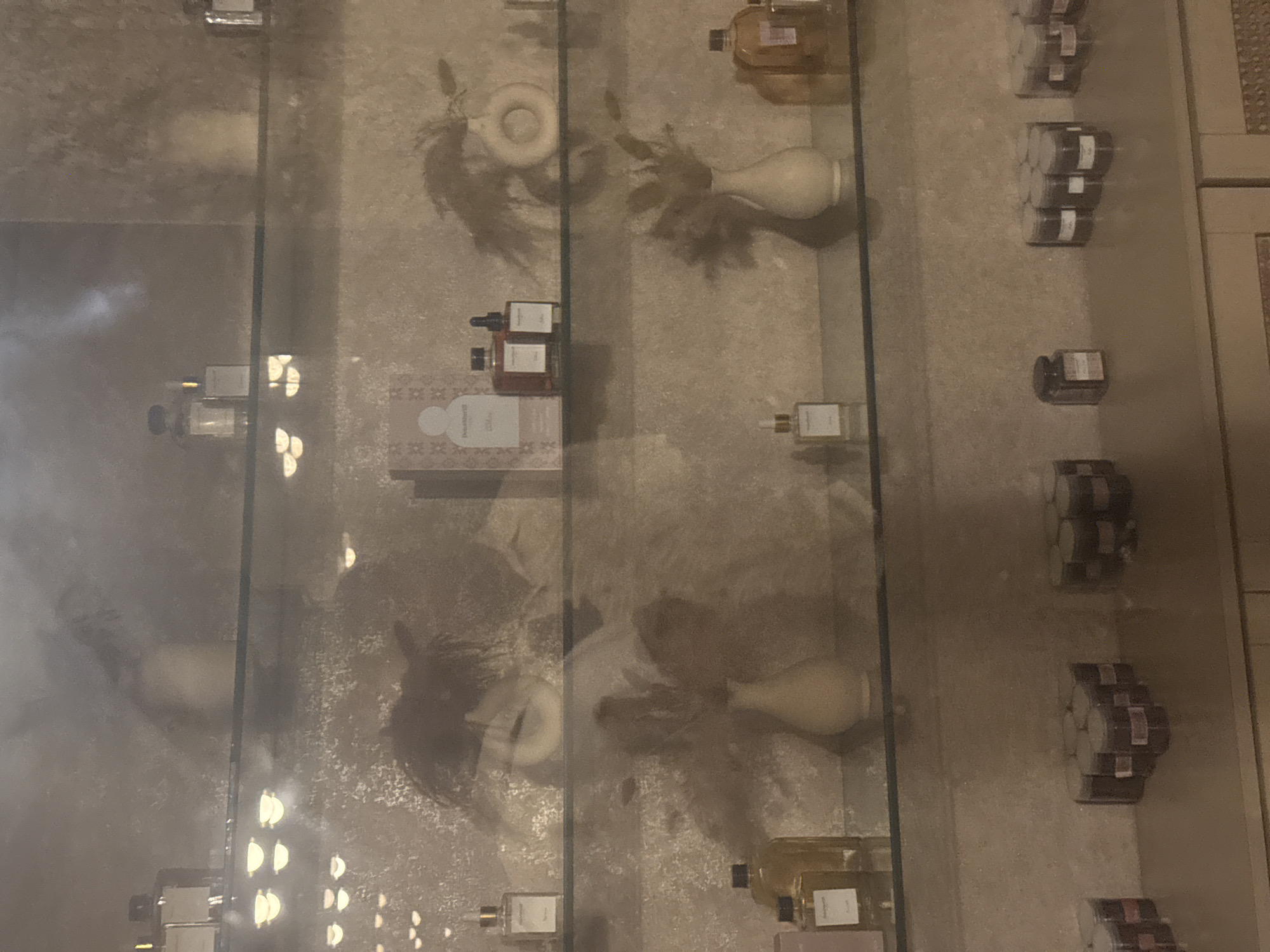 A dimly lit interior scene showcases shelves filled with various perfume bottles and small jars, likely in a retail setting. The display is illuminated by soft lighting, casting reflections on the glass shelves and creating shadowy silhouettes of people on the textured wall behind the products. The overall ambiance suggests a boutique or a high-end store. This scene is located in Abuja, Nigeria.