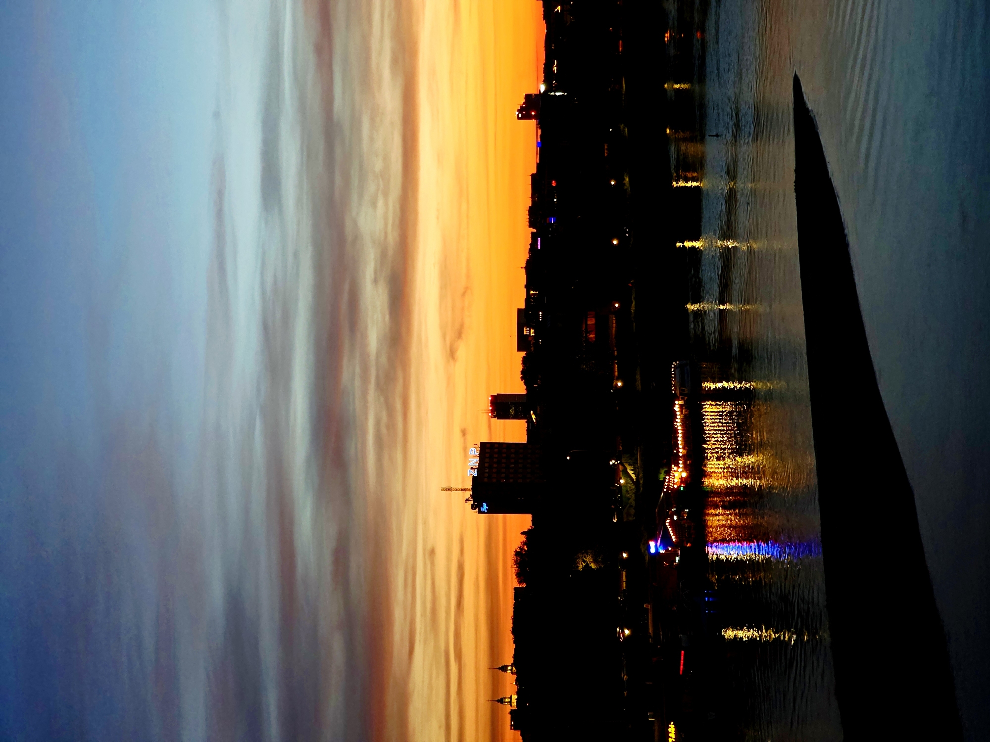The image shows a sunset over the Vistula River in Warsaw, Poland. The sky is filled with vibrant orange and yellow hues, with streaks of blue and purple clouds. The city skyline is silhouetted against the colorful sky, with various buildings and structures visible. The river reflects the lights from the city, creating a shimmering effect on the water. There are no people or animals clearly visible in the image. The scene appears to be a quiet evening in the city.