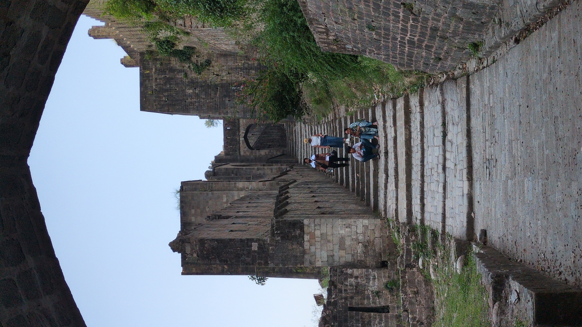 Captured from below and rotated, the image showcases a section of the Golconda Fort, situated in Kaswara, India. Dominating the foreground are weathered, stone walls which enclose a steep stairway. Several people are on the steps. Three appear to be seated on the steps. A man is standing and is wearing a white shirt, blue jeans, and a dark jacket. Another man stands on the steps facing away from the viewer and he is wearing a white shirt, a brown jacket, and dark pants. The vegetation is overgrown. A clear sky is overhead.