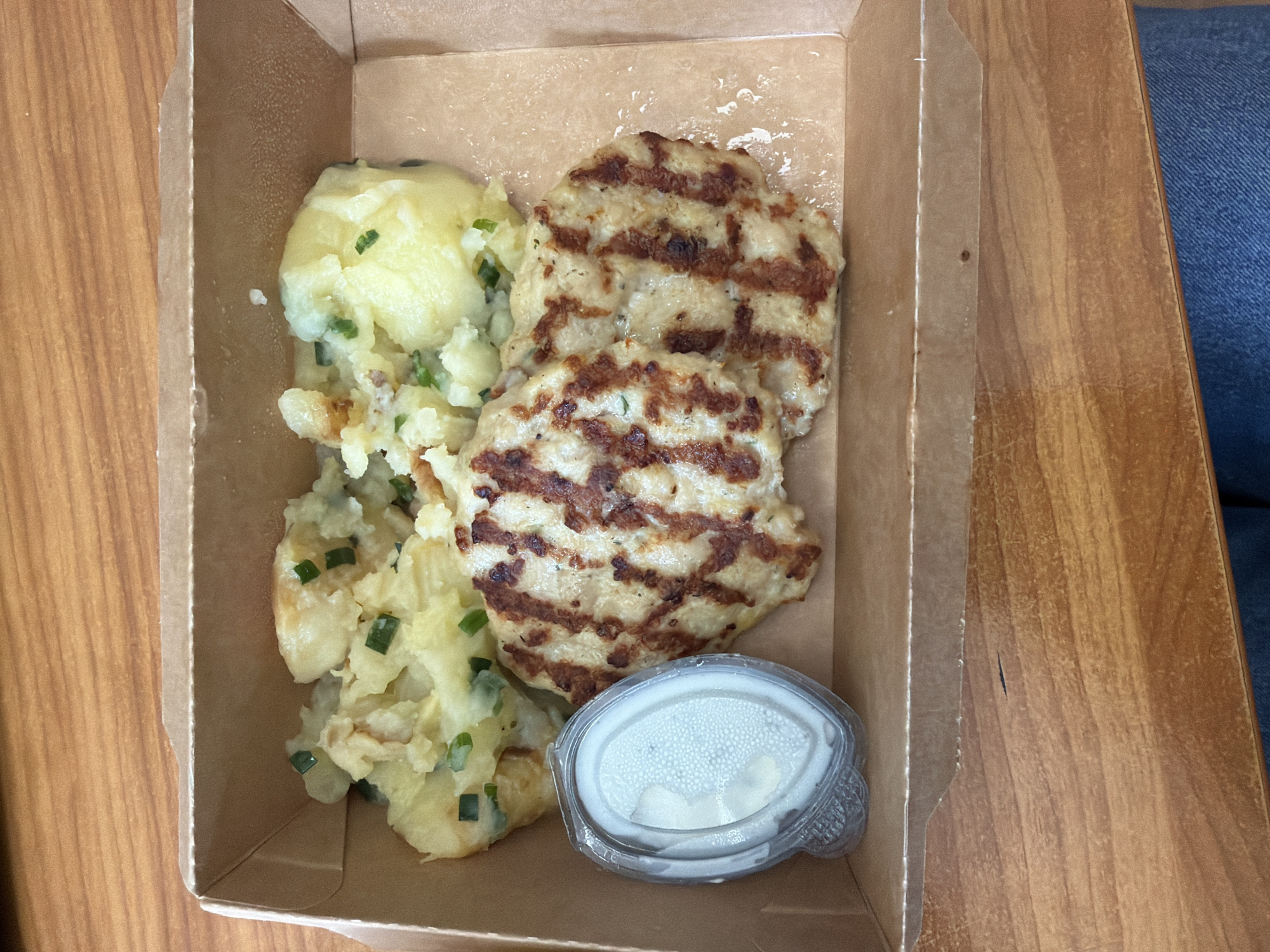 A top-down shot shows a cardboard container holding a meal consisting of mashed potatoes with green herbs, two grilled patties, and a small container of white sauce. The meal is situated on a wooden surface, possibly a table. Part of a denim-clad leg is visible in the upper right corner. The photo appears to be taken indoors. The image does not contain enough information to determine the location in Sofia, Bulgaria.