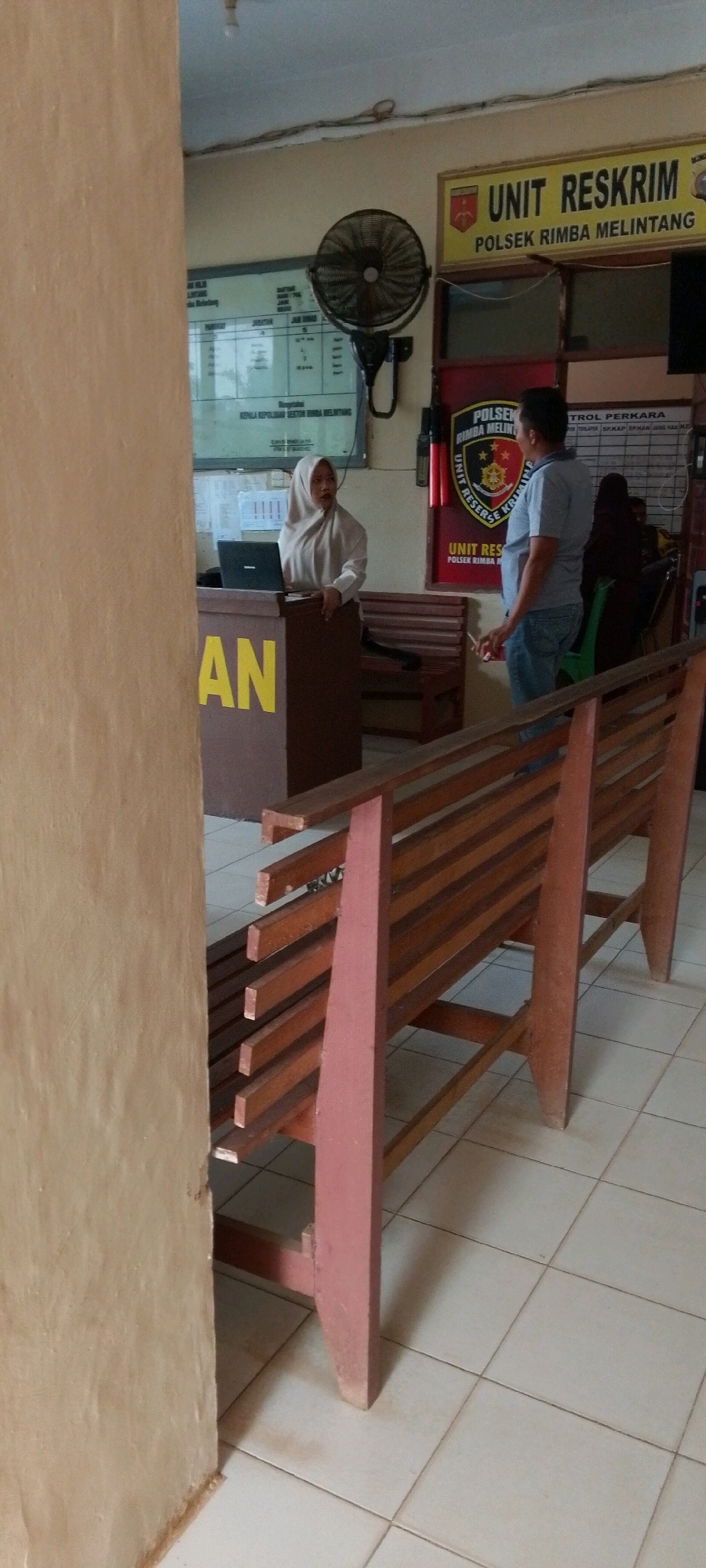 This is a full-shot photograph of the interior of what appears to be a police station lobby in Unknown, Indonesia. In the foreground, a brown wooden bench with red-painted legs is positioned diagonally across the frame. Behind the bench, a woman in a light-colored hijab stands behind a dark brown podium with "AN" in large yellow letters on the front. She is looking to her right and appears to be interacting with someone off-camera. A laptop is open on the podium in front of her. To the right of the podium, a man wearing a blue t-shirt and jeans stands facing the woman, also looking towards her. Behind them, on the wall, is a yellow sign that reads "UNIT RESKRIM POLSEK RIMBA MELINTANG". A circular fan is mounted on the wall above and to the left of the sign. There are other signs and what appears to be a bulletin board on the wall behind the man. Two more wooden benches are visible further back in the room. The floor is tiled in a light color.