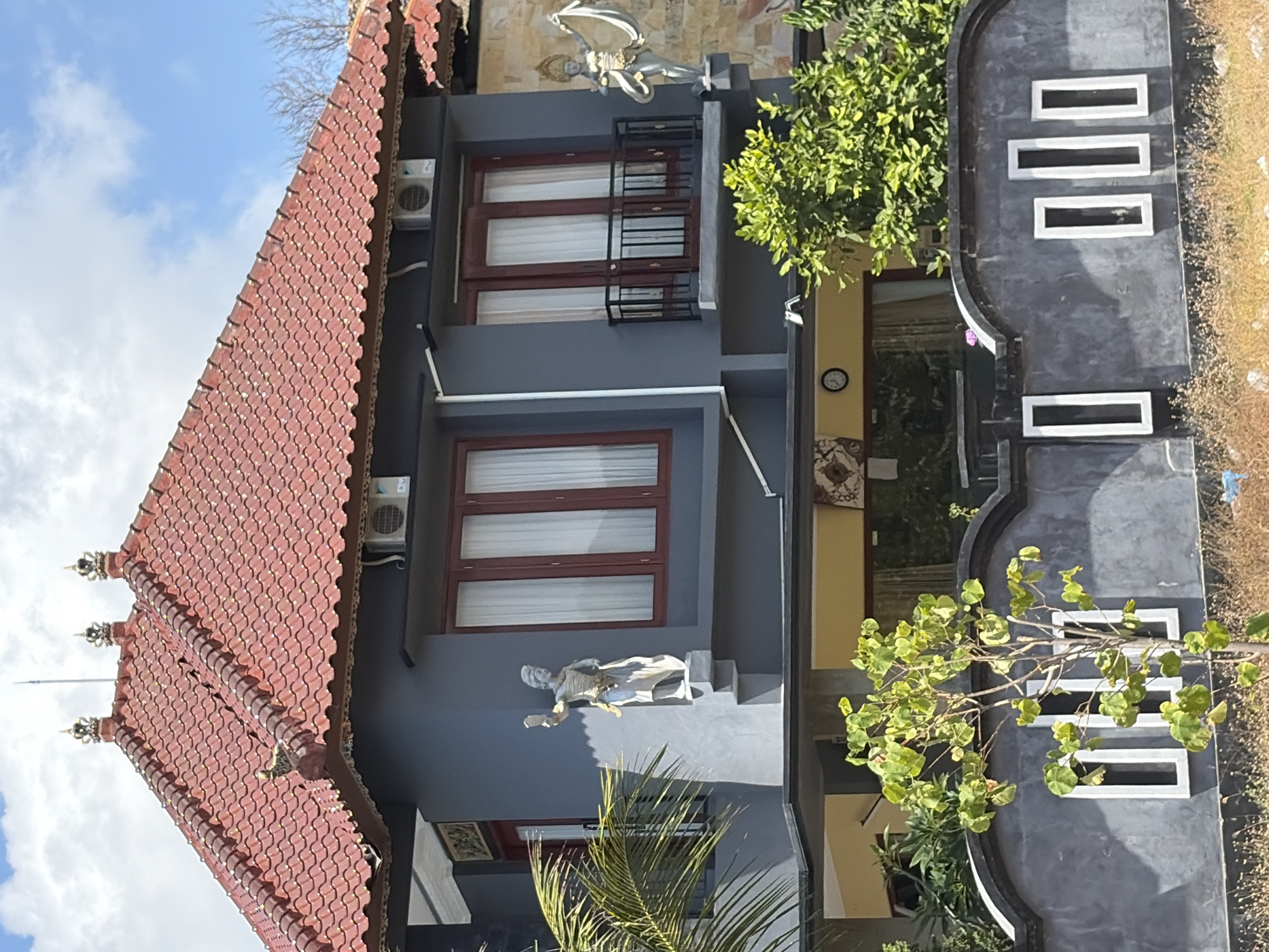 A two-story building with a red tiled roof is pictured on a bright, sunny day in Nusa Penida, Indonesia. The building's facade is painted gray, with two upper-story windows on the left and two on the right. The windows have dark red frames and white curtains. A small balcony with black railings is situated between the right-side windows. On the left side of the building, a statue of a person in a traditional outfit is mounted on the wall. A similar statue is visible on the right side of the building, slightly further back and partially obscured by greenery. A palm frond extends into the frame from the left. In the foreground, a dark gray wall with decorative rectangular cutouts runs across the bottom of the image. A small tree with green leaves grows in front of the wall, and dry grass and small debris are visible at the very bottom. The sky above is blue with scattered white clouds.