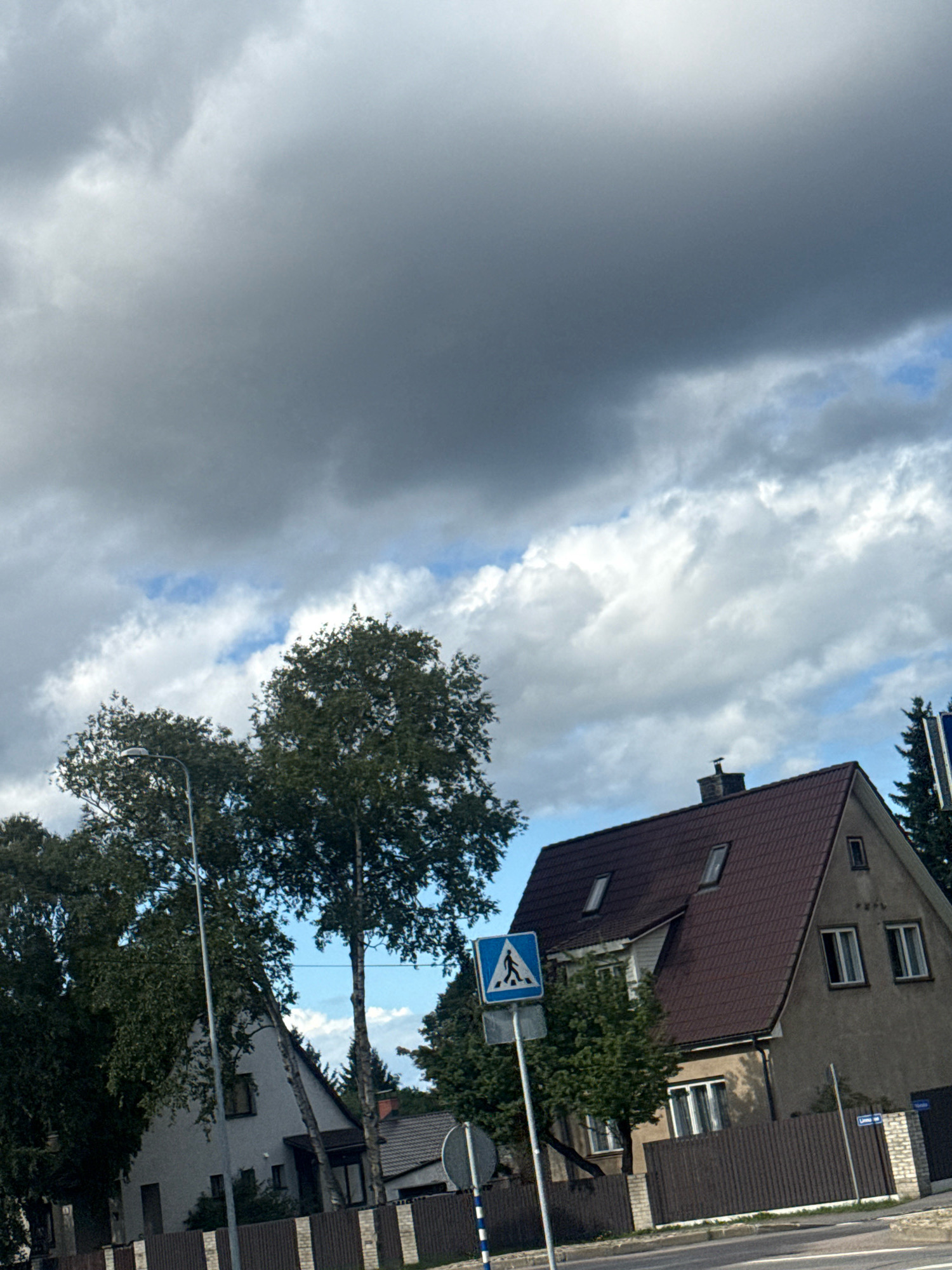 This is a street-level, eye-level, medium shot of a residential area in Tallinn, Estonia, under a cloudy sky. The image captures a juxtaposition of suburban architecture and natural elements. In the foreground, a dark brown wooden fence runs horizontally across the frame, bordering a street. To the left, a tall, slender streetlight stands prominently. Behind the fence, a light-colored, two-story house with a dark red, sloped roof is visible. This house features several windows and a chimney. Adjacent to it, a cluster of mature trees with dark green foliage adds a natural element to the scene. To the right of this house, another building with a similar dark red roof and lighter-colored walls is partially visible. A blue pedestrian crossing sign, featuring a white silhouette of a person walking, is mounted on a pole to the left of the main house. The sky is filled with dramatic, dark gray clouds, interspersed with patches of lighter, white clouds and hints of blue sky. The overall impression is a slightly overcast day in a quiet residential neighborhood.