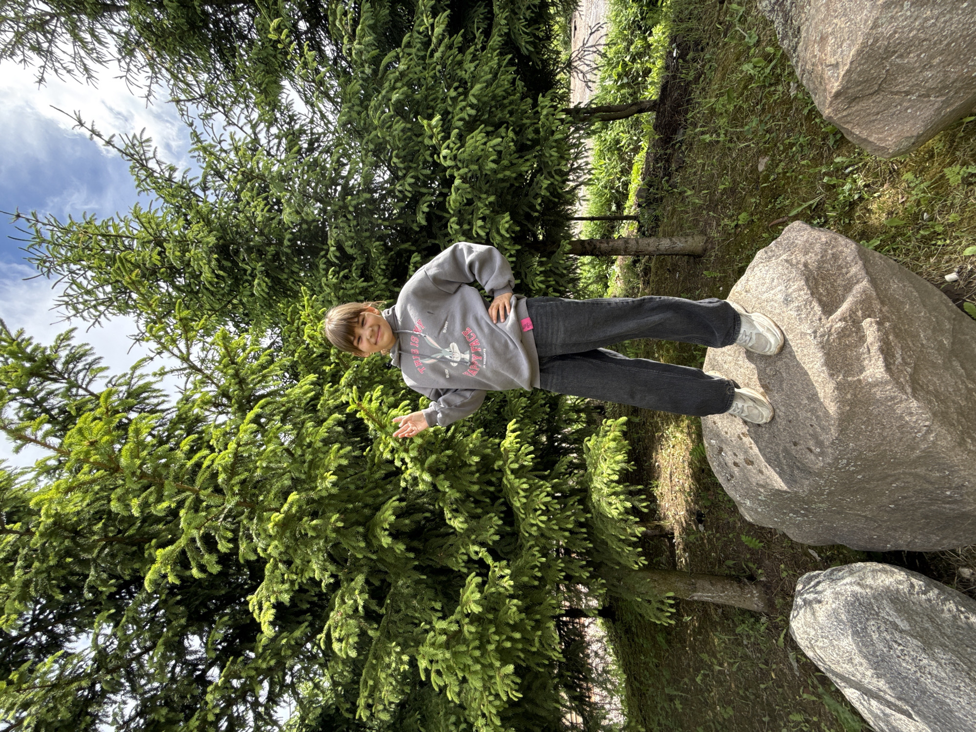 A young person stands on a rock in what appears to be a park-like setting, possibly in Rusanovo, Russia. The person is wearing a gray hoodie and dark-colored jeans with white sneakers. They are smiling and holding up two fingers in a gesture. The environment includes evergreen trees, grass, and several large rocks. The sky is visible in the upper left, indicating an outdoor location.