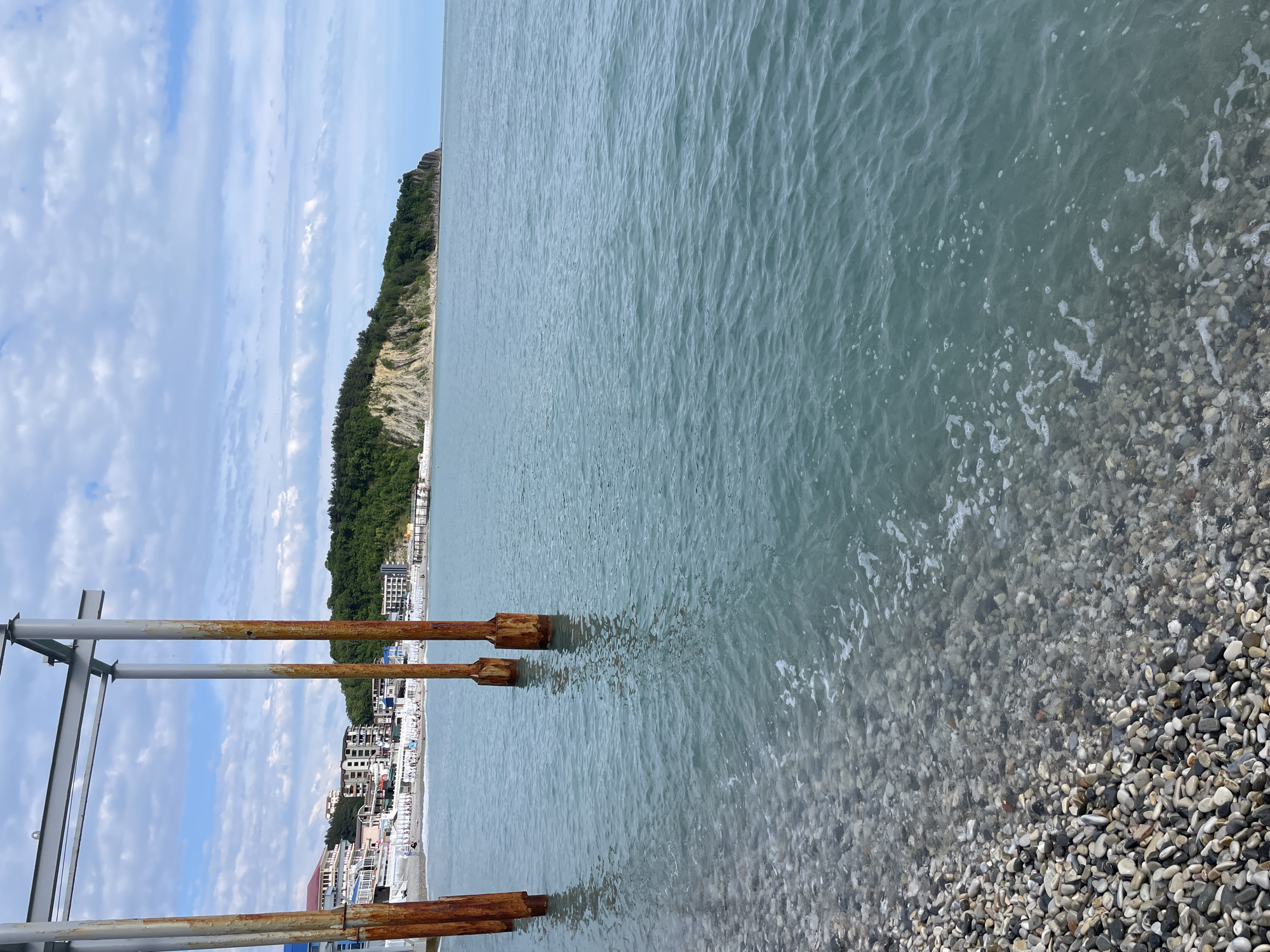 The image depicts a beach scene in Olginka, Russia. The perspective is from a pier or structure with rusty metal supports extending into the water. A pebble beach transitions into a calm, light turquoise sea. In the background, a coastal town is visible with buildings, a beach area, and a cliff covered in vegetation. The sky is partly cloudy. There are no people visible in the immediate foreground.