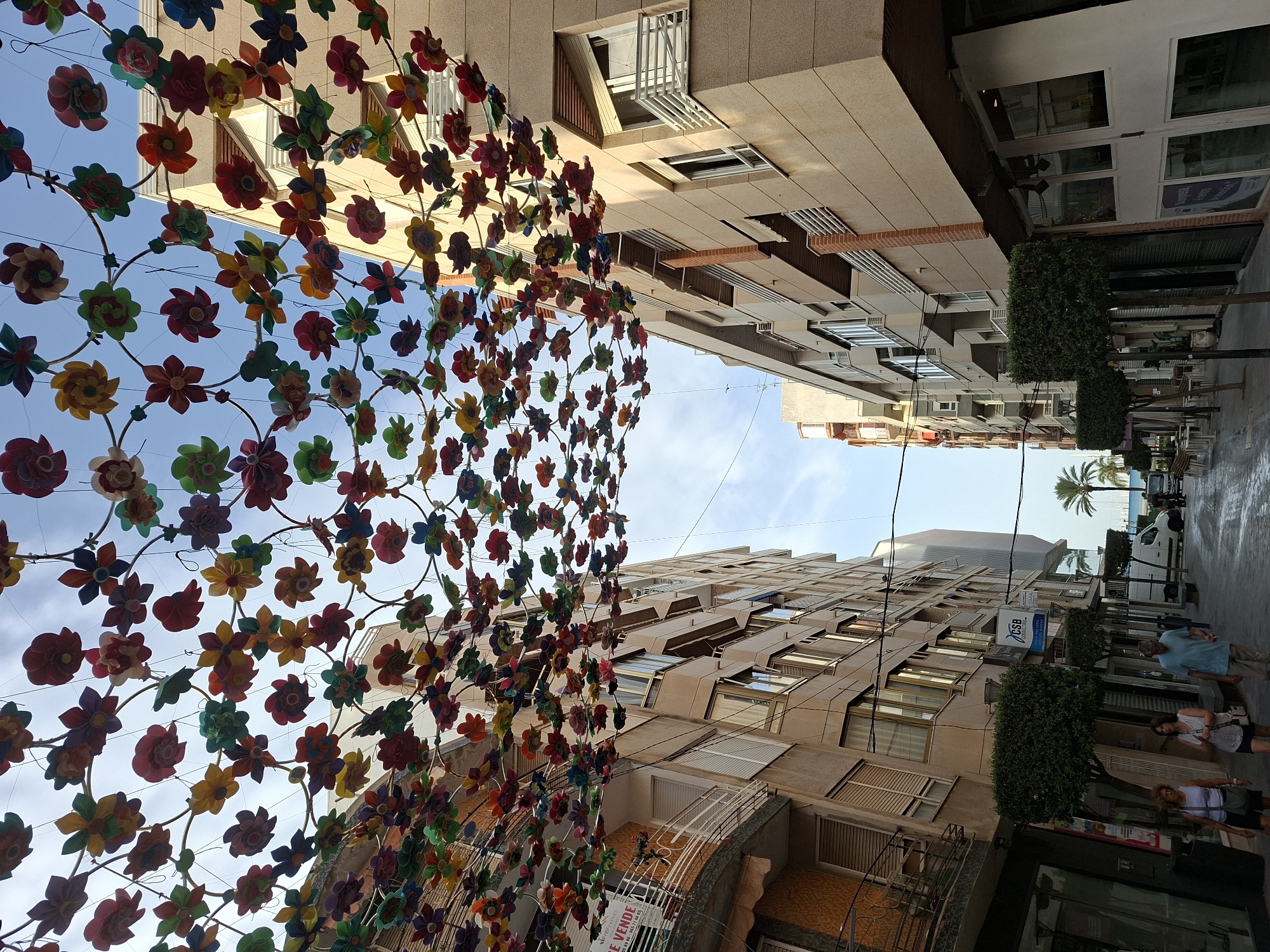 The image captures a narrow street in Torrevieja, Spain, adorned with a canopy of colorful, artificial flowers strung across the sky. The street is lined with multi-story buildings, characterized by their light-colored facades, balconies with white railings, and numerous windows. On the left side, a building features a "SE VENDE" (FOR SALE) sign. The street itself appears to be paved, with some areas wet, suggesting recent rain or cleaning.
Several people are visible walking along the street. Two women are walking together towards the foreground, followed by a man. Further down the street, a white van and other vehicles are parked, and more people can be seen in the distance. The scene is framed by manicured, square-shaped bushes planted along the sidewalk. Palm trees are visible in the background, hinting at the coastal location. The sky above is partly cloudy.