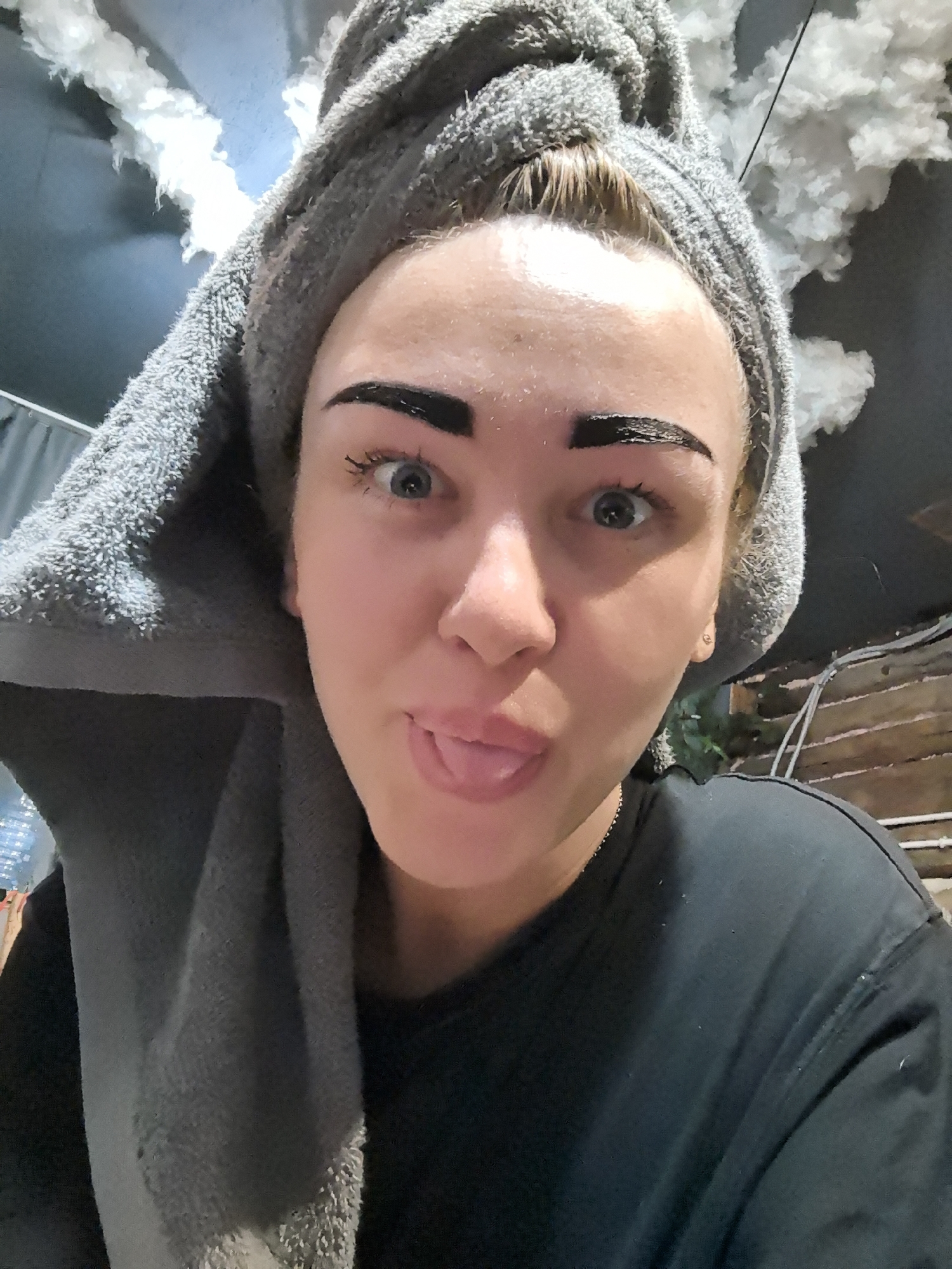 A fair-skinned woman with dark eyebrows, covered in a dark substance, and a grey towel wrapped around her head is sticking out her tongue. She is wearing a dark T-shirt. The background includes what appears to be cloud-like decorations on a ceiling. There is no evidence to indicate the location of Noviy, Russia.