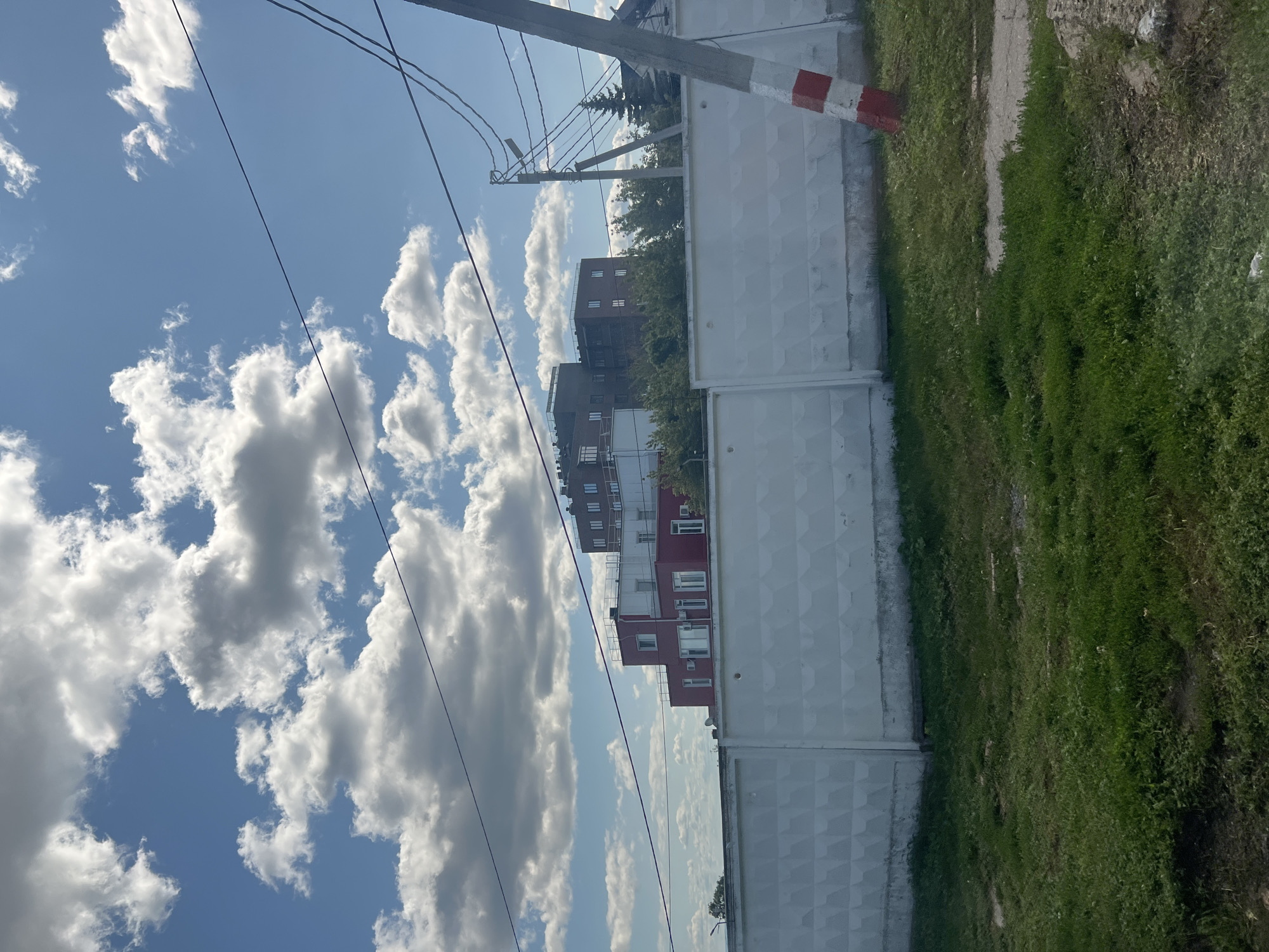 The image shows a view of buildings and a wall in Ulyanovsk, Russia. The buildings are modern, with multiple stories and windows. One building is red and white, while another is dark gray. A white, textured concrete wall runs across the foreground, obscuring the view of the buildings behind it. Green grass and some dirt are visible in front of the wall. Overhead, there are power lines and a bright blue sky with fluffy white clouds. A red and white striped pole is visible on the right side of the image, likely part of utility infrastructure. No people are present in the image.